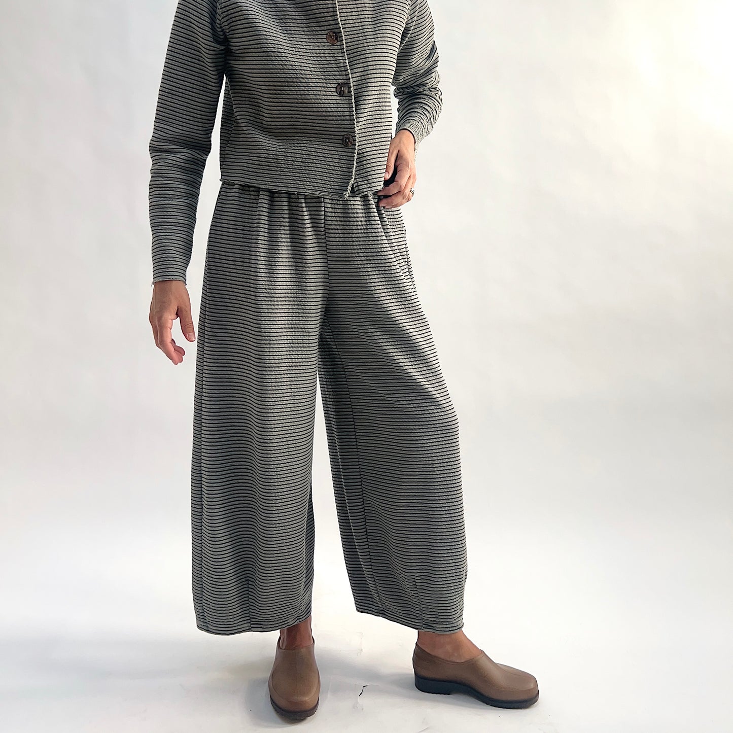 Full front view of model wearing squirrel and black fine horizontal stripe wide-leg cropped pants, hands on hips showing the elastic waist and dart shaping, styled with the matching striped asymmetrical cardigan and taupe slip-on shoes against a white background.
