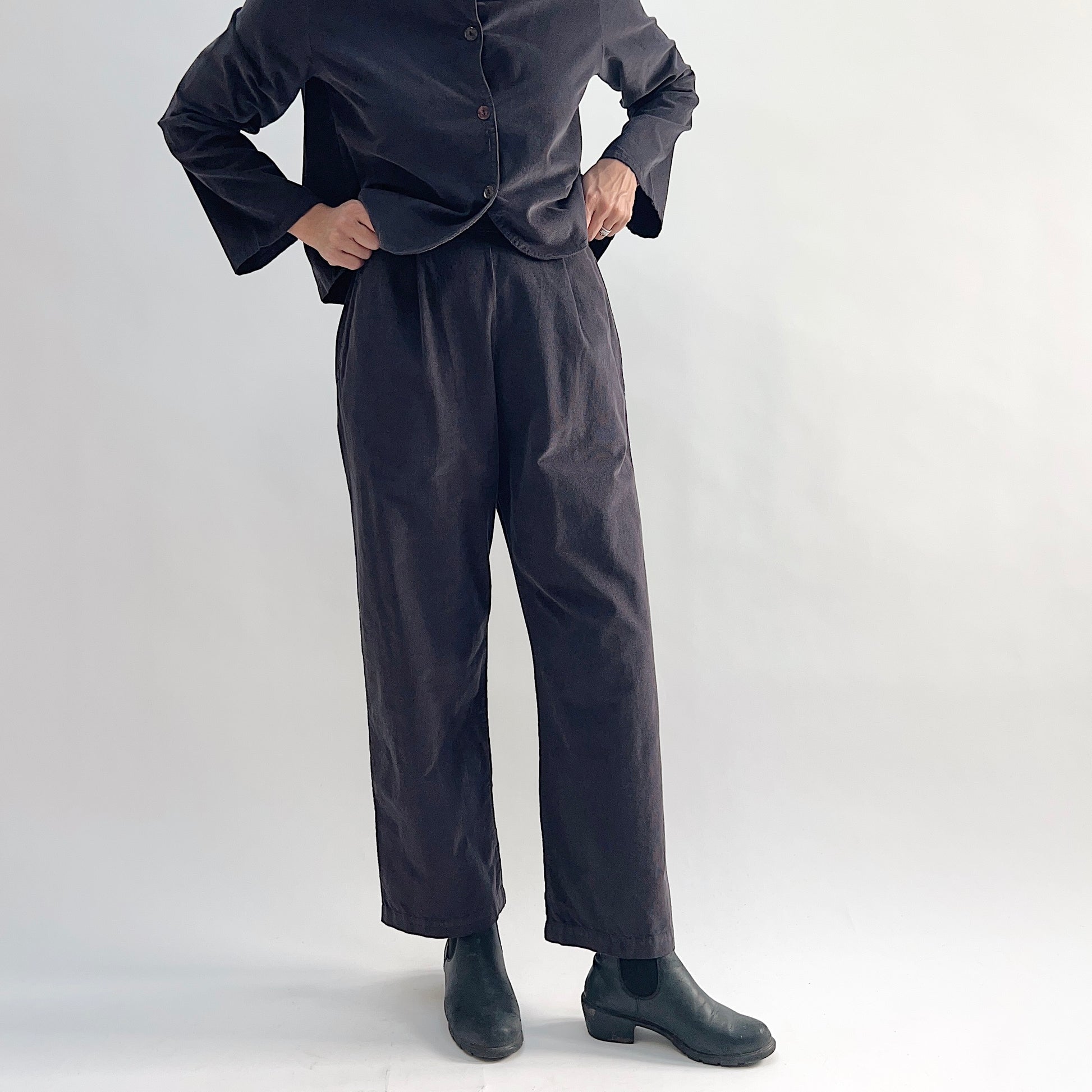 Model wearing the Bryn Walker Corduroy Pleated Pants in Fig, cropped front view showing flat front with pleats, elastic waistband, and tapered straight leg in deep fig pinwheel corduroy, styled with matching Standing Collar Crop Jacket and black Chelsea boots
