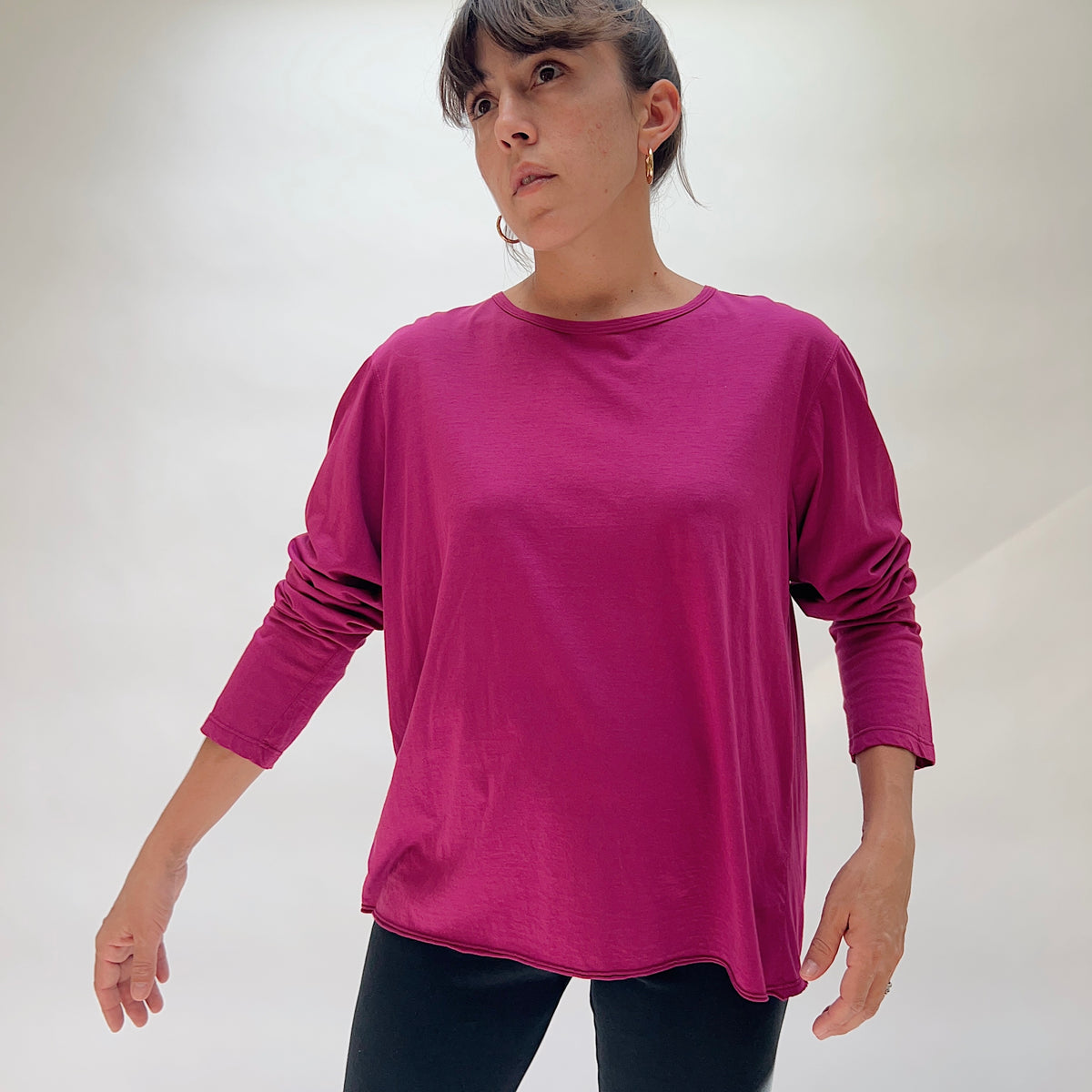 Model wearing the North Star Base Tissue Cotton Long Sleeve Agnes Tee in Raspberry, shown from a slight angle — a relaxed boxy fit crew neck tee in rich magenta raspberry tissue cotton with long sleeves and a curved lettuce-edge hem, paired with dark trousers
