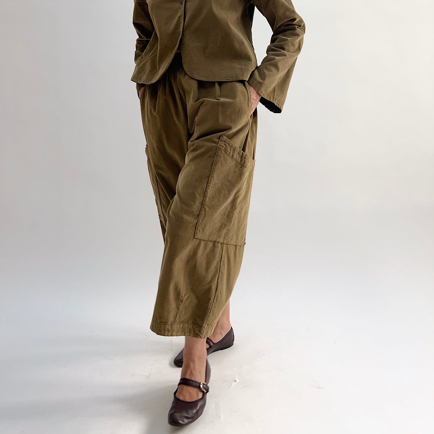 Model with hand on hip wearing the Bryn Walker Corduroy Pasha Pants in Partridge, front view showing side leg patch pockets, elastic waistband, and olive pinwheel corduroy, paired with matching Audrey Jacket and dark Mary Jane flats
