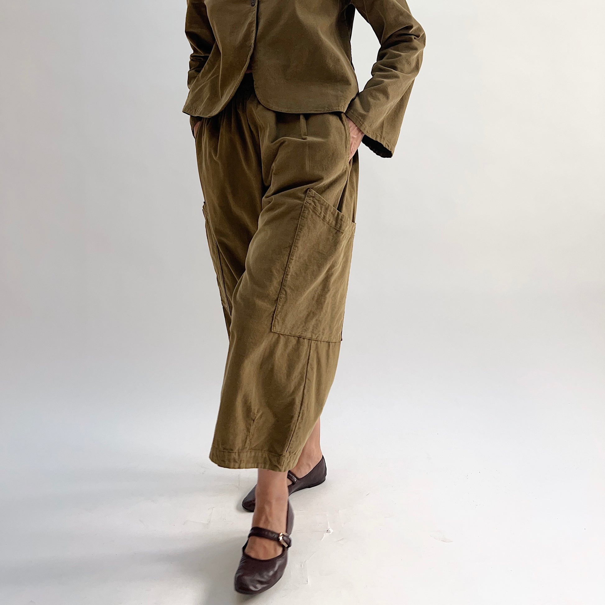 Model with hand on hip wearing the Bryn Walker Corduroy Pasha Pants in Partridge, front view showing side leg patch pockets, elastic waistband, and olive pinwheel corduroy, paired with matching Audrey Jacket and dark Mary Jane flats
