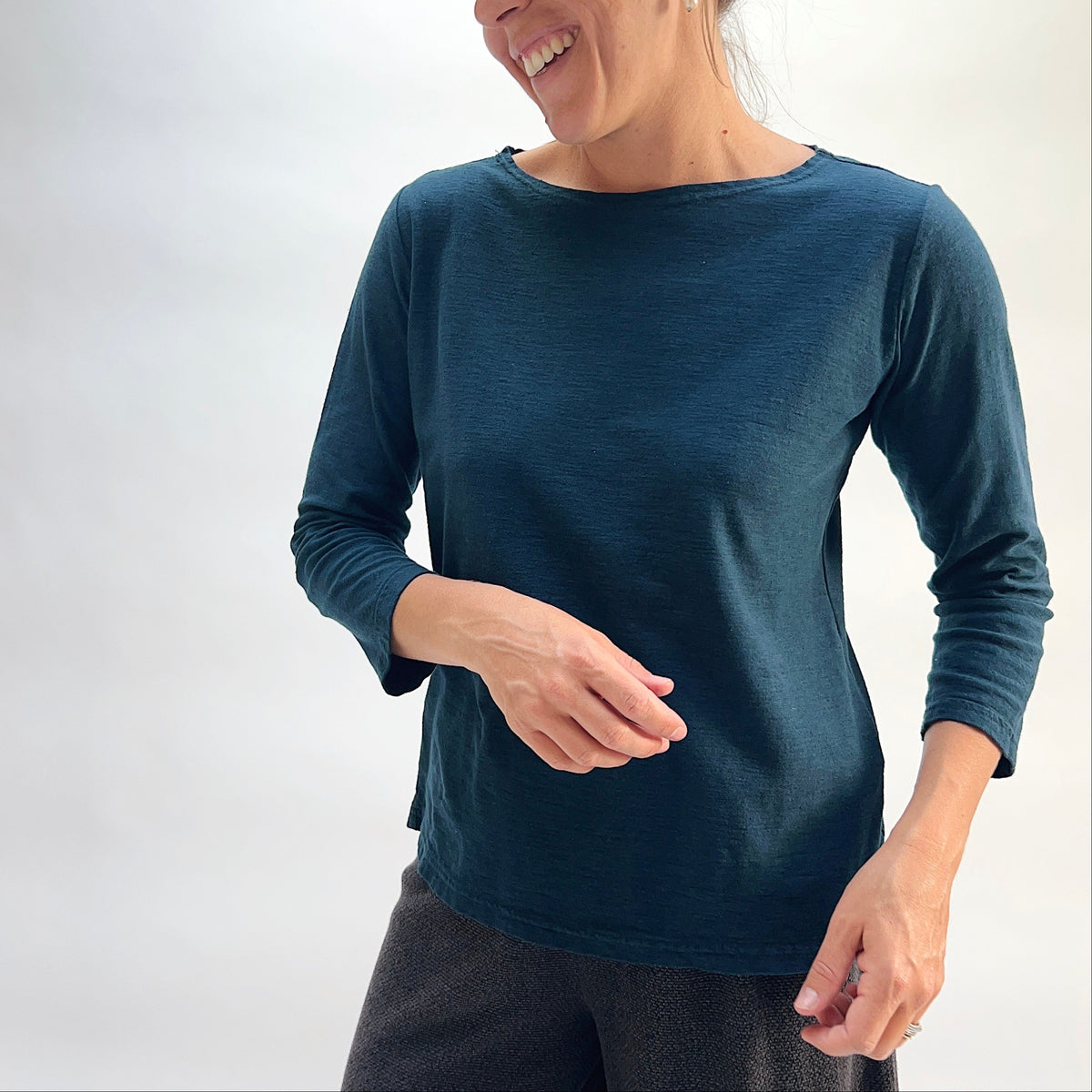 Model wearing the Cut Loose 3/4 Sleeve Boatneck Top in Etan, smiling three-quarter front view, showing boatneck neckline and relaxed straight hem of deep teal cotton-linen top with 3/4 sleeves, styled with black textured pants
