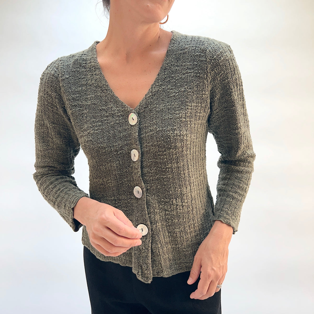 Model wearing the Cut Loose Puff Sleeve Cardigan in Kelp, close-up front view fastening bottom button, showing large natural shell button detail, V-neckline, and marled knit texture of olive green cardigan with scalloped hem, styled with black pants
