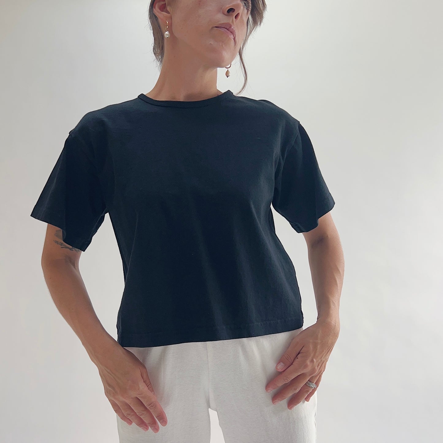 Full front view of model wearing Eleven Stitch black crop crew tee with hands on hips, showing crew neckline, short sleeves, relaxed boxy fit, slightly cropped length, and black color, styled with white wide-leg pants

