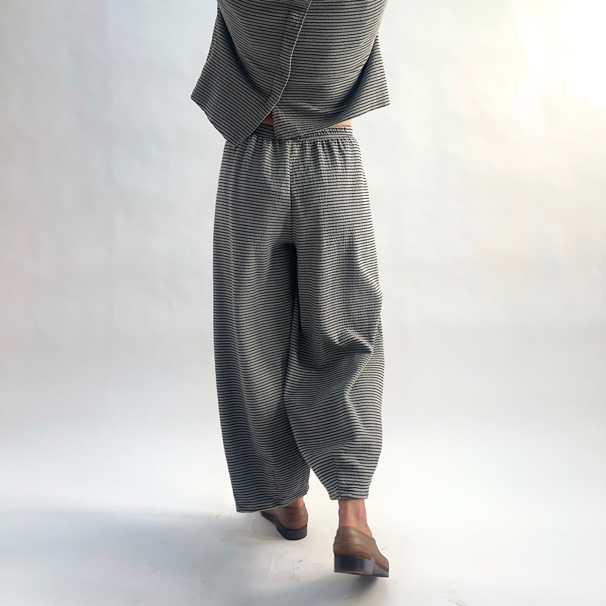 Back view of model wearing squirrel and black fine horizontal stripe wide-leg cropped pants with elastic waist, showing the relaxed wide-leg silhouette and crinkle-texture fabric, styled with the matching striped cardigan and taupe slip-on shoes.
