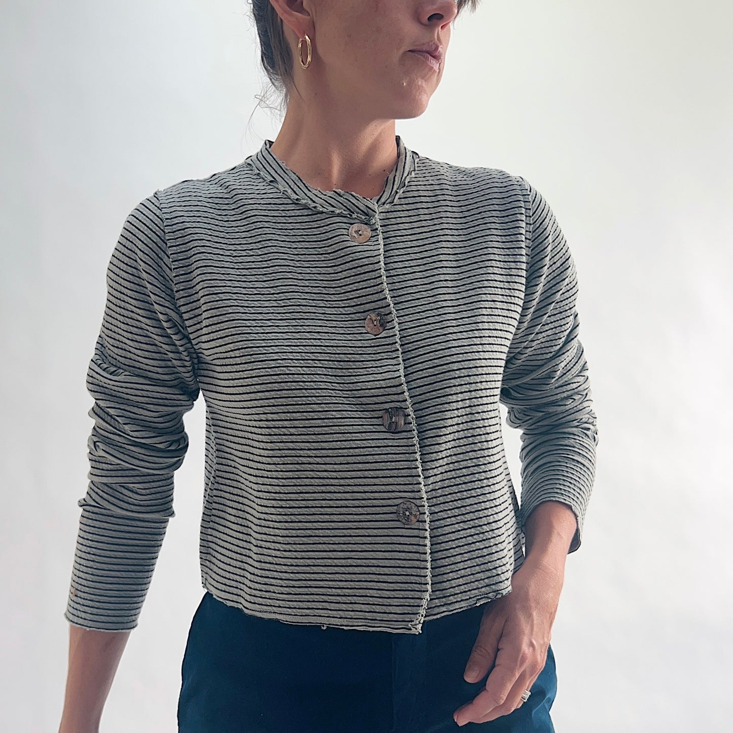 Close front view of model wearing a squirrel and black fine horizontal stripe cropped cardigan, showing the asymmetrical button closure, band neckline, and crinkle-texture stripe detail, styled with dark teal wide-leg pants.
