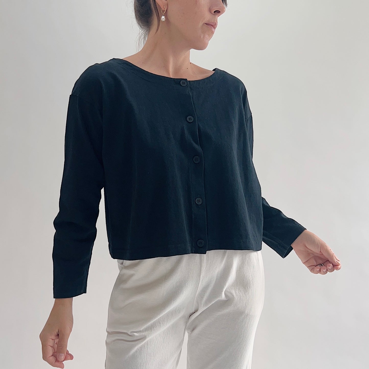 Three-quarter front view of model wearing Pacific Cotton black long sleeve button front shirt, showing wide scoop neckline, five-button center placket, long sleeves, boxy cropped fit, and black color