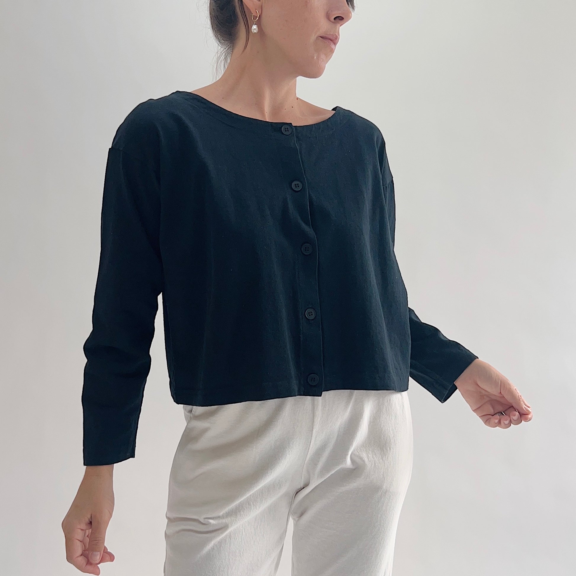 Three-quarter front view of model wearing Pacific Cotton black long sleeve button front shirt, showing wide scoop neckline, five-button center placket, long sleeves, boxy cropped fit, and black color