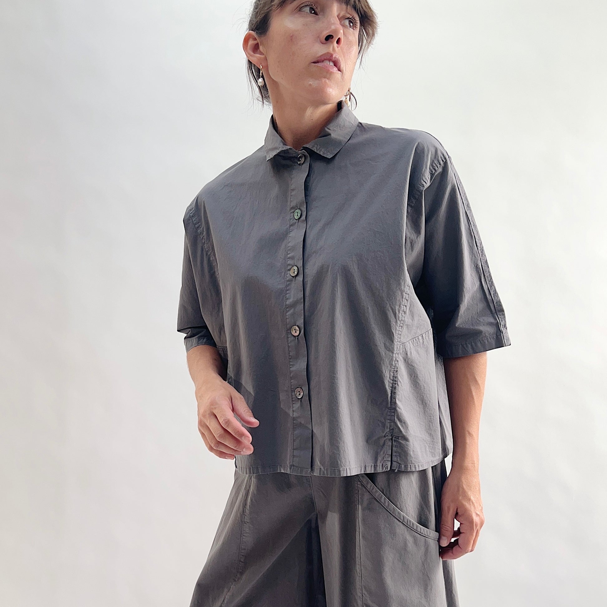 Full front view of model wearing Eleven Stitch eiffel tower camp shirt looking up, showing point collar, dark shell buttons, short sleeves, patch pocket, slightly cropped boxy fit, side slits, and deep slate grey color, styled with matching eiffel tower one pocket pant