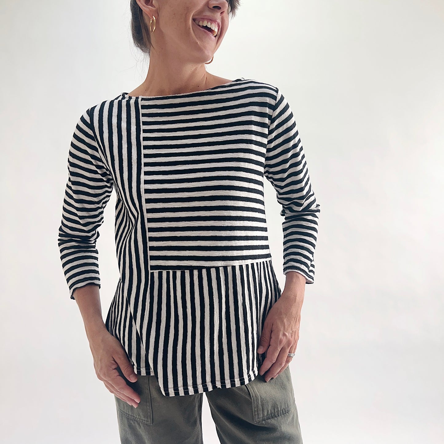 Smiling front view of model wearing a black and white patchwork stripe A-line boatneck top, showing the mixed horizontal and vertical stripe panel construction and flared A-line silhouette.
