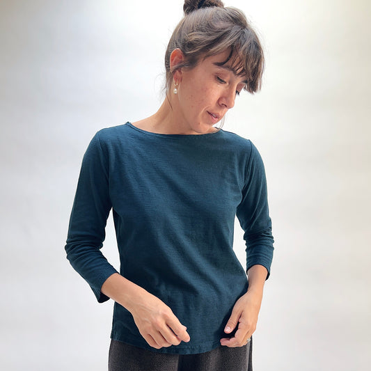 Model wearing the Cut Loose 3/4 Sleeve Boatneck Top in Etan, close-up three-quarter view looking down, deep teal cotton-linen boatneck top with 3/4 sleeves and subtle slub texture, styled with black textured pants
