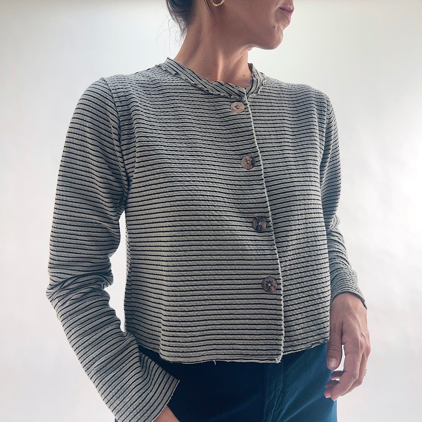 Three-quarter front view of model wearing a squirrel and black fine horizontal stripe cropped cardigan with asymmetrical button closure and long sleeves, looking to the side, showing the relaxed cropped silhouette and crinkle-texture fabric, styled with dark teal wide-leg pants.
