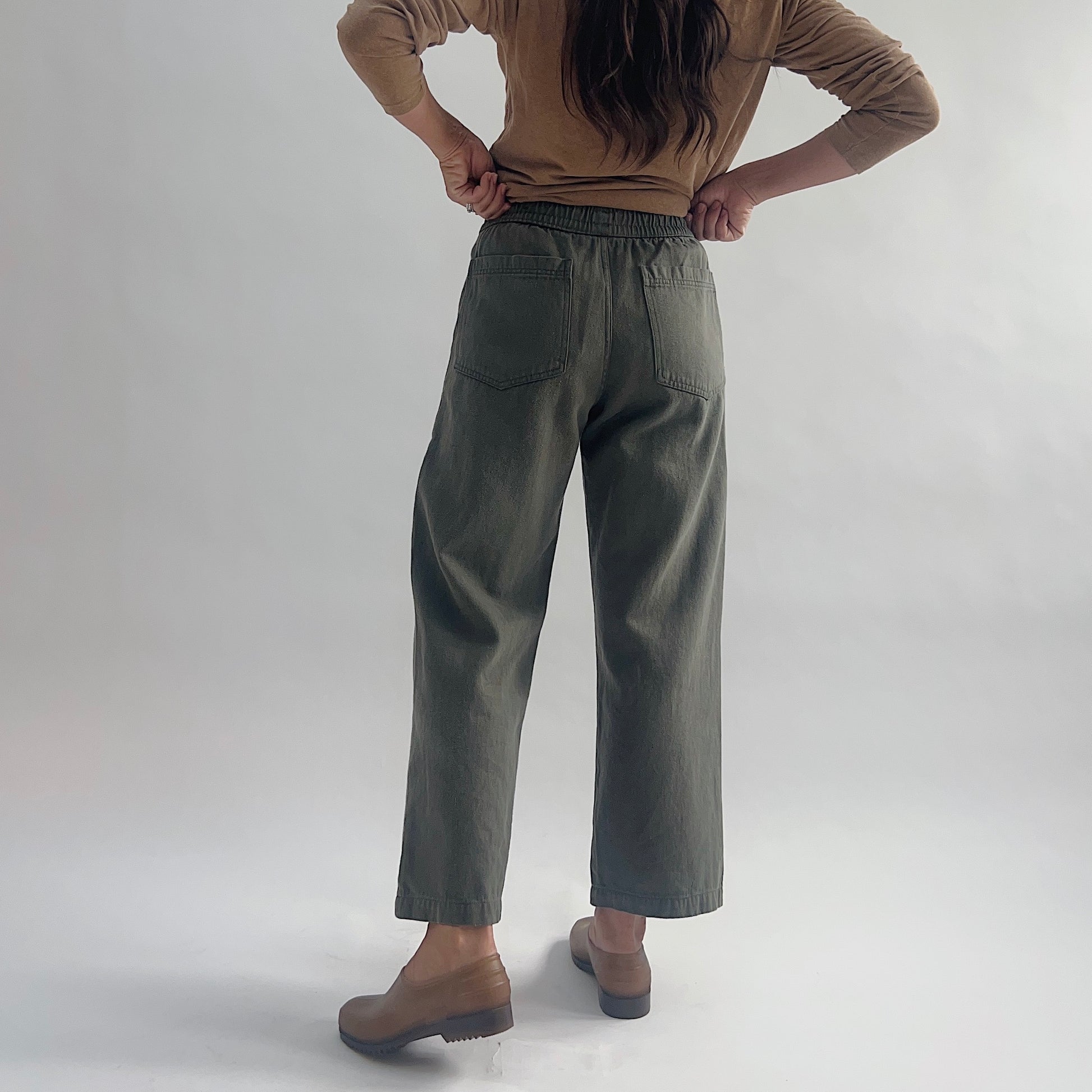 Back view of model wearing olive green wide-leg pull-on pants with two back patch pockets and elastic waist, styled with a tan long-sleeve top and brown clogs.

