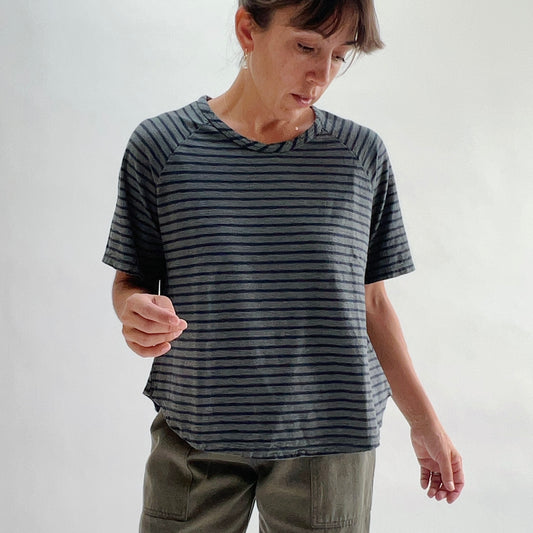 Front view of model looking down wearing an anthracite and navy striped boxy raglan tee with a relaxed fit and boat neckline, paired with olive green pants.
