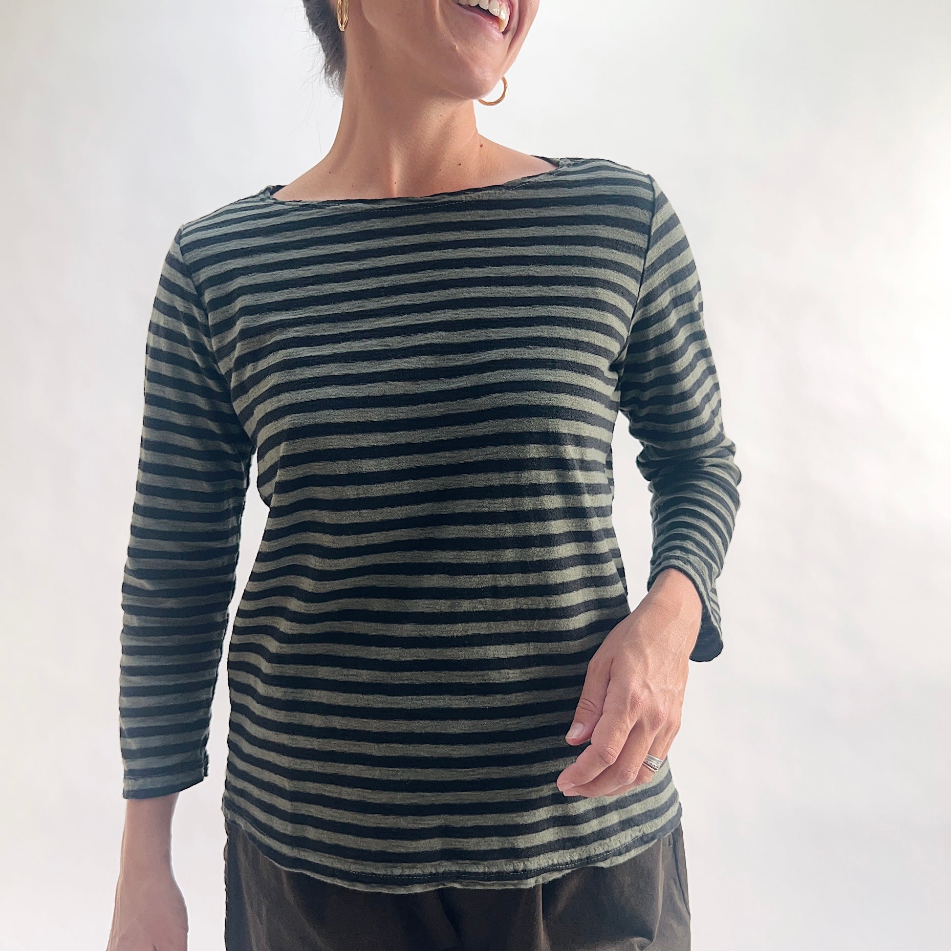 Smiling close front view of model wearing a kelp green and black horizontal stripe boatneck tee with 3/4 sleeves, hand on hip, showing the wide boatneck neckline and linen-blend texture, styled with dark charcoal pants.
