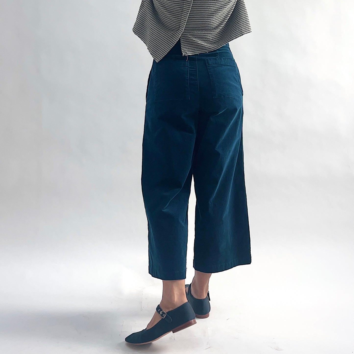 Back view of model wearing deep teal mini cord straight crop trousers, showing the back welt pockets, wide straight leg silhouette, and cropped hem, styled with the matching white and black stripe asymmetrical cardigan and teal mary jane flats against a white background.
