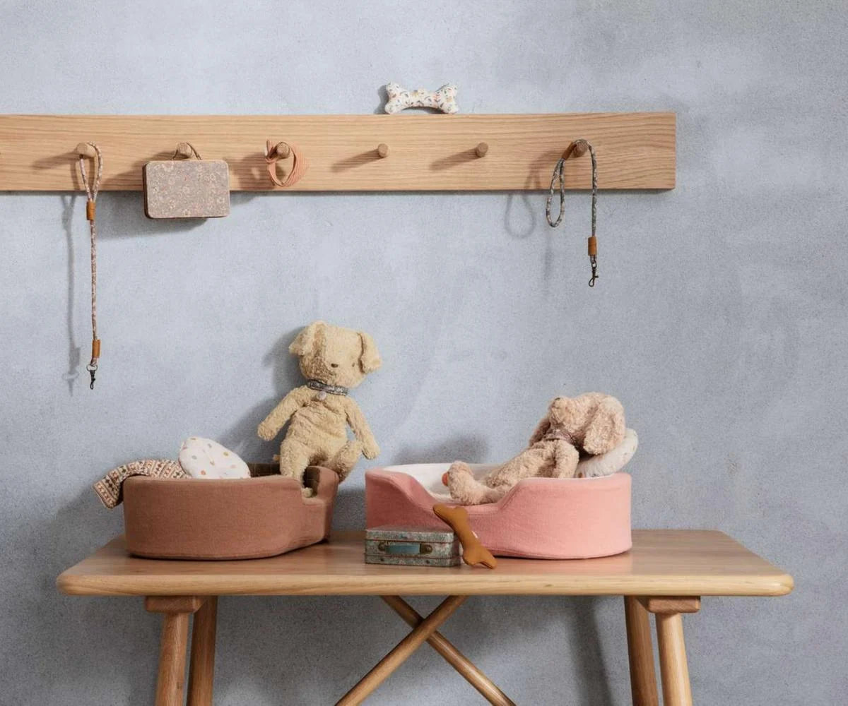 Maileg Plush Dog in Soft Sand styled in a Maileg dog-themed scene — two soft plush dogs sitting in terracotta and pink oval dog beds on a wooden bench, with miniature leashes, a toy bone, and a suitcase, against a grey wall with a wooden peg rail
