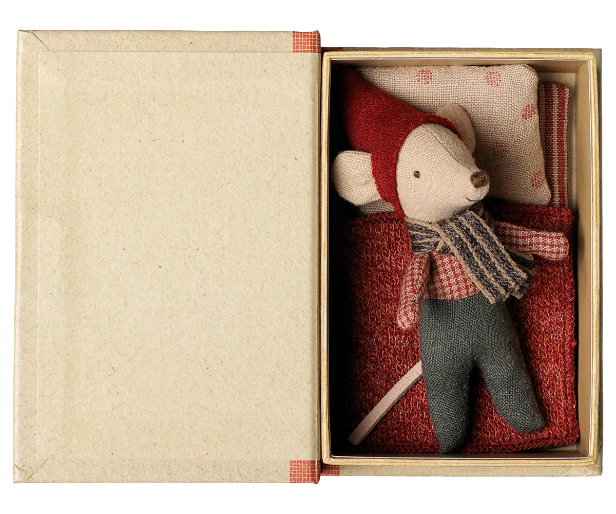 Maileg Christmas baby mouse wearing a red hat and striped scarf, resting on red bedding with a patterned pillow inside an open book box.

