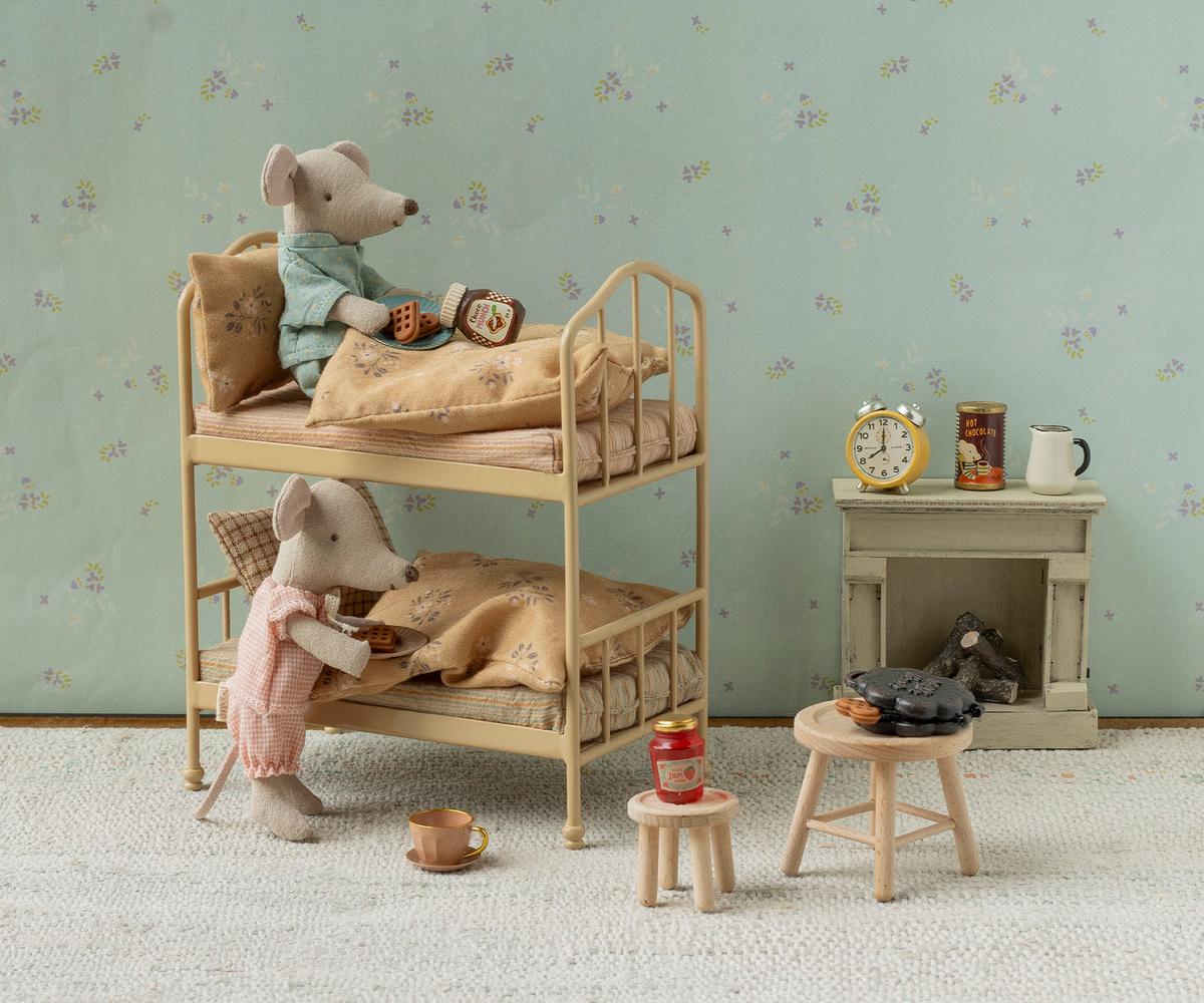 Maileg Bunk Bed in Wheat — lifestyle scene of two Maileg mouse dolls in a cosy bedroom, one in a teal top resting on the top bunk with a waffle and honey jar, one in a pink romper standing at the lower bunk holding a tea tray, beside a miniature fireplace and wooden stools

