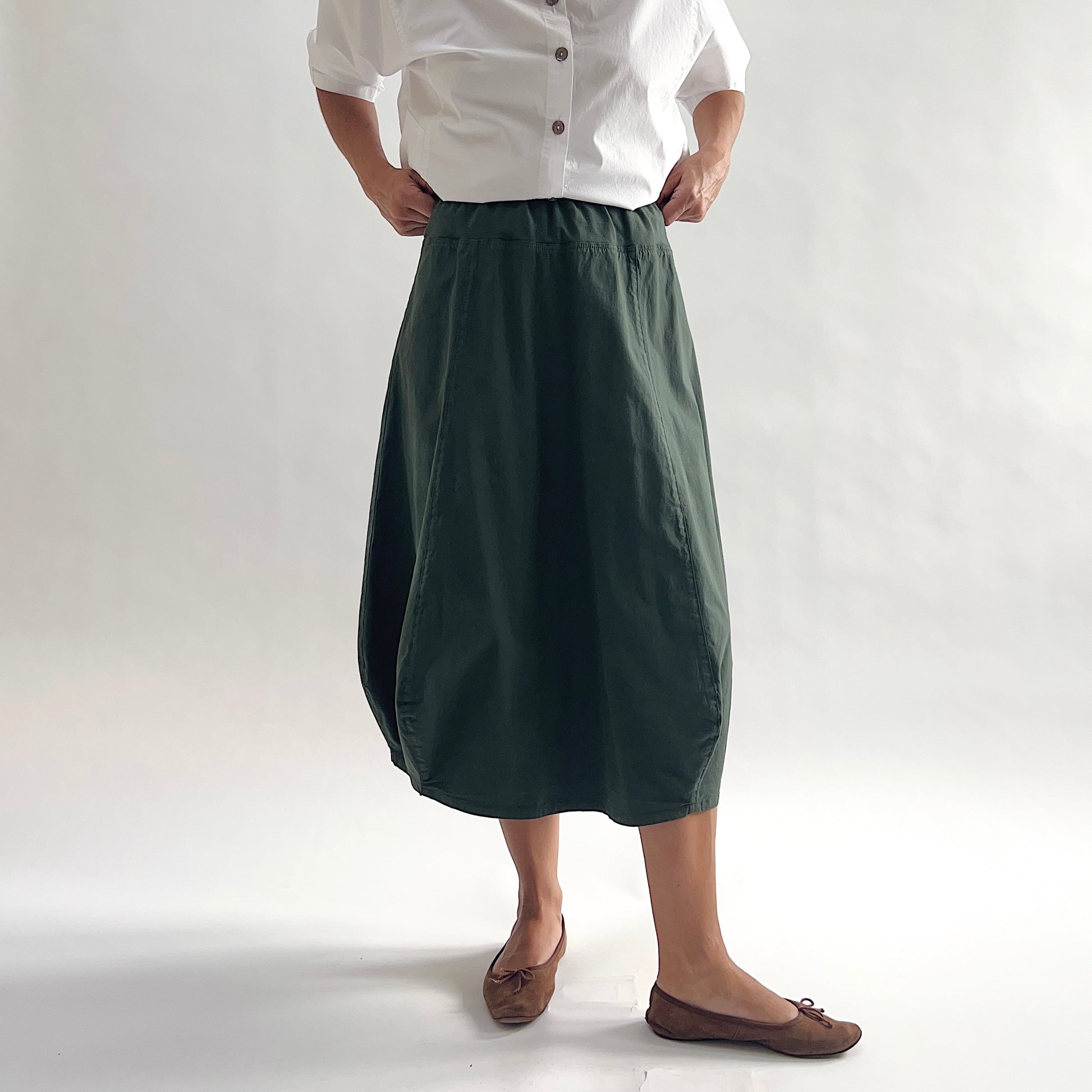 Full front view of model wearing Eleven Stitch konbu rib yoke skirt with hands on waist, showing elastic knit waistband, curved seam paneling, sculptural bubble hem, below-the-knee length, and deep forest green color, styled with white chalk camp shirt and brown ballet flats