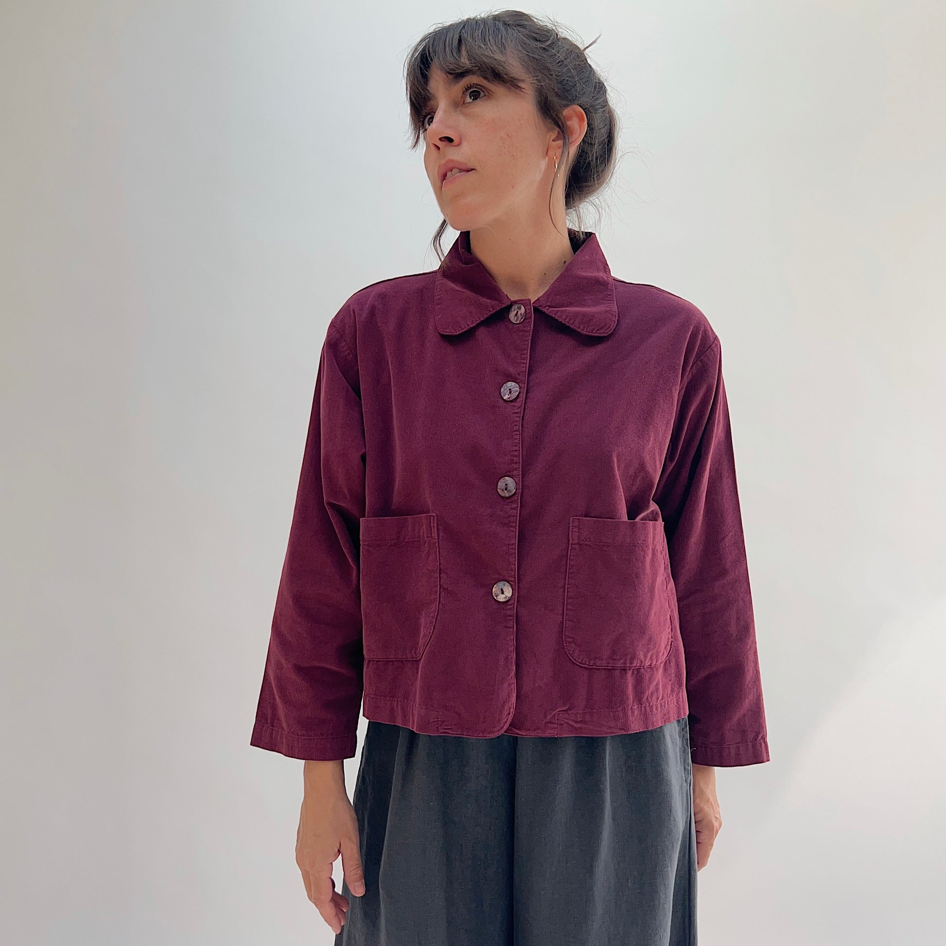 Model wearing the Bryn Walker Corduroy Audrey Jacket in Rhone, full front view showing rounded collar, shell button closure, patch pockets, and relaxed cropped silhouette, paired with grey skirt
