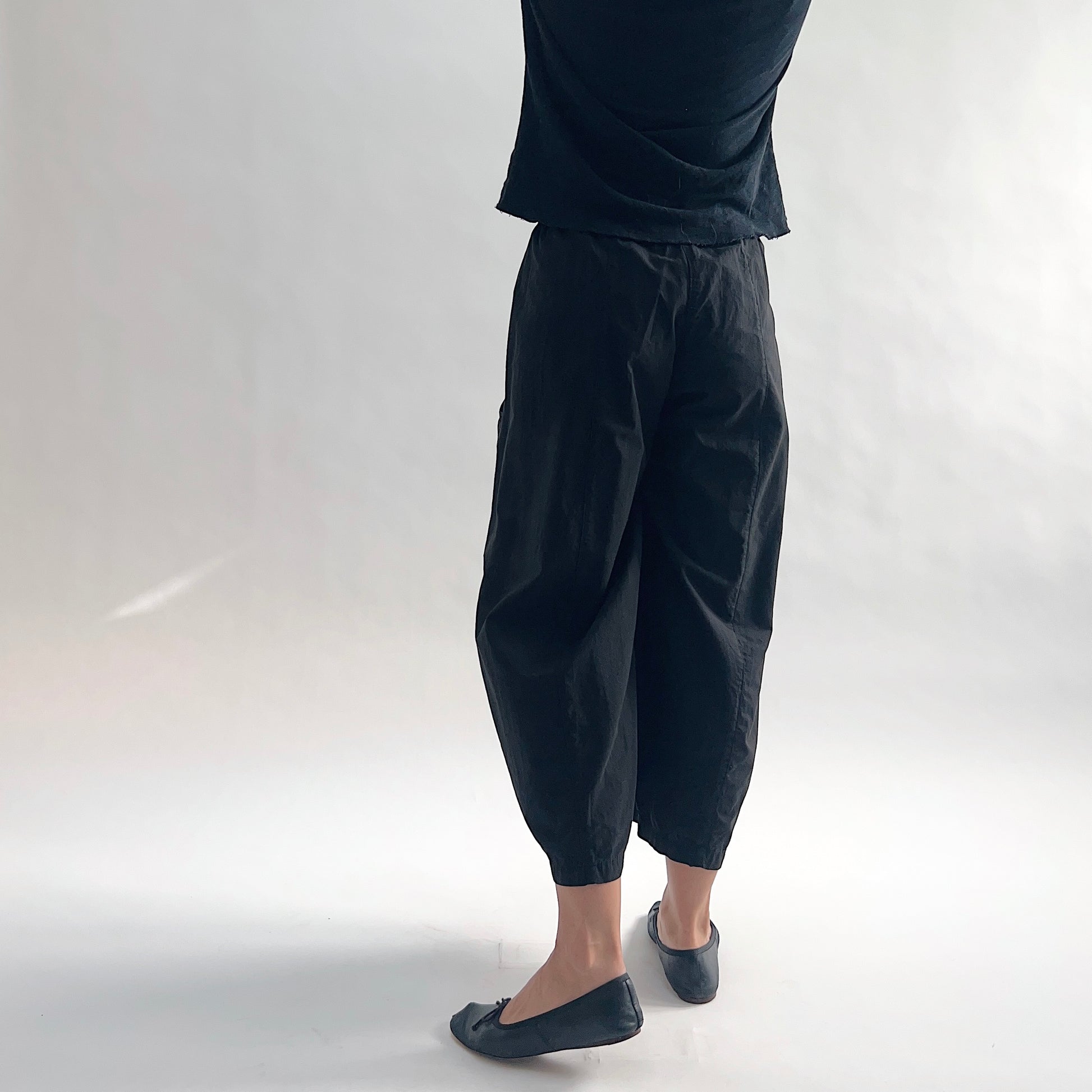 Back three-quarter view of model wearing Eleven Stitch black one pocket pant, showing elastic waistband, wide curved-seam cropped leg, gathered elastic hem, and black color, styled with black top and black ballet flats