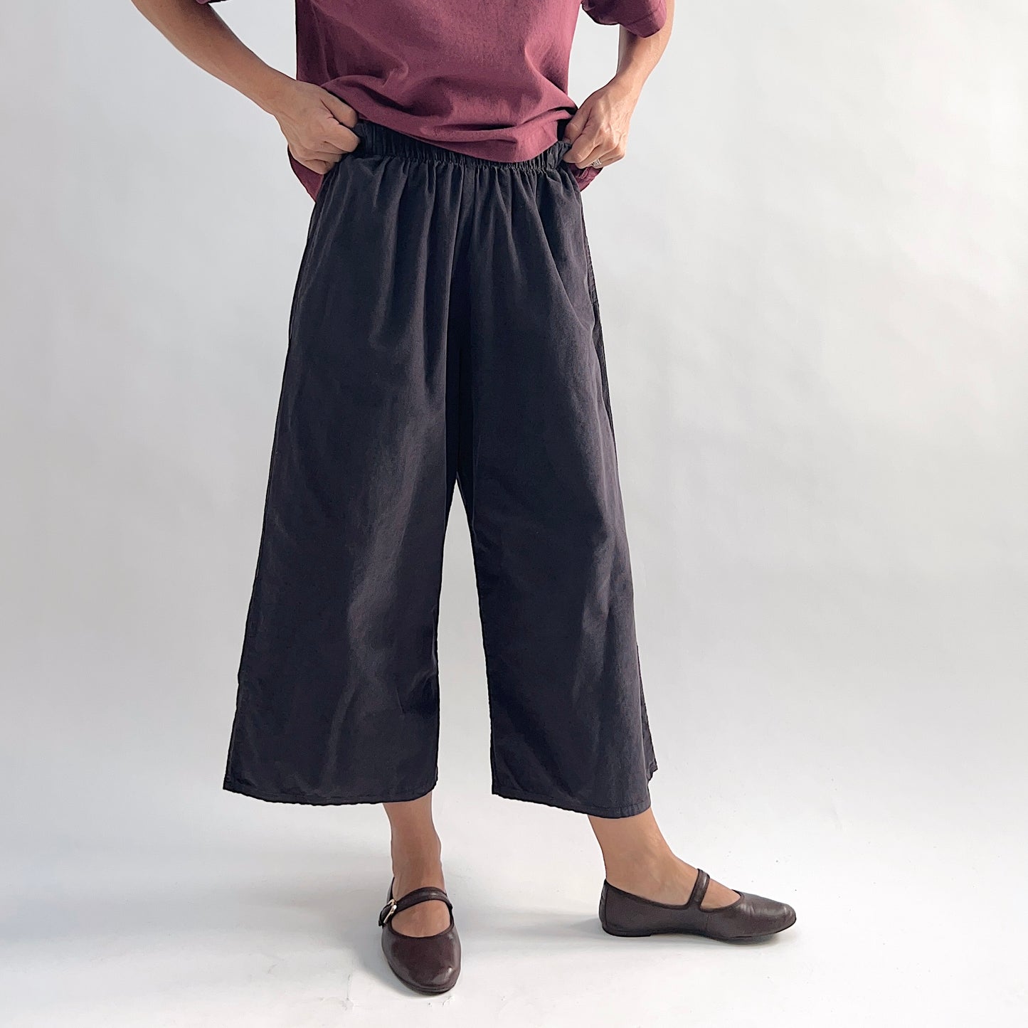 Model lifting top to show waistband of the Bryn Walker Corduroy Flood Pants in Fig, full front view highlighting fully elastic gathered waistband, wide culotte-style leg silhouette, mid-calf flood length, and deep fig pinwheel corduroy, styled with burgundy Double Cotton Tee and brown Mary Jane flats
