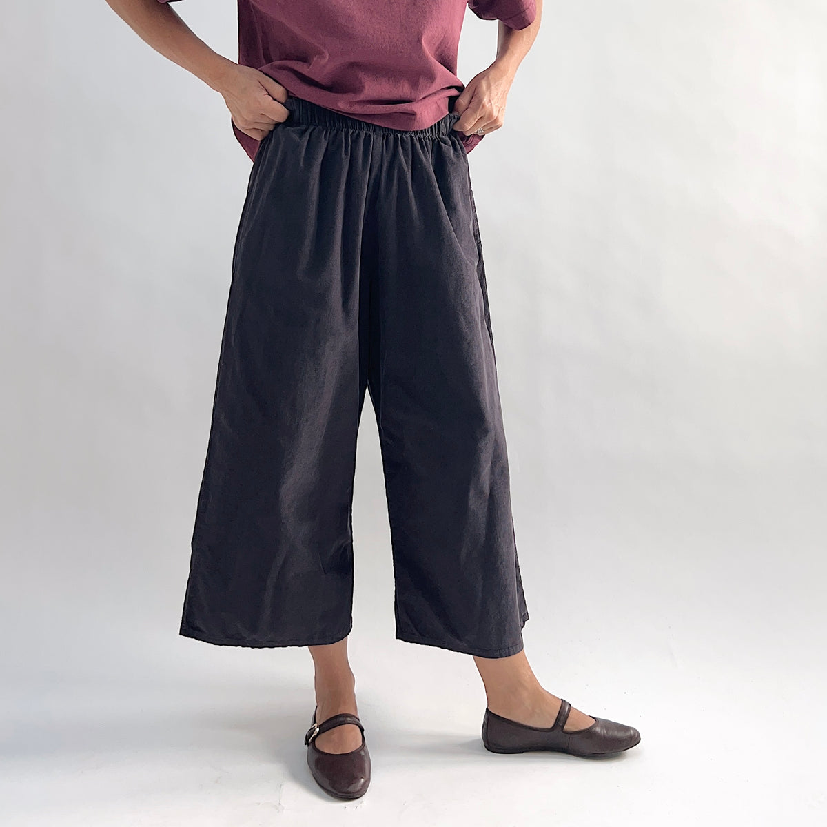 Model lifting top to show waistband of the Bryn Walker Corduroy Flood Pants in Fig, full front view highlighting fully elastic gathered waistband, wide culotte-style leg silhouette, mid-calf flood length, and deep fig pinwheel corduroy, styled with burgundy Double Cotton Tee and brown Mary Jane flats
