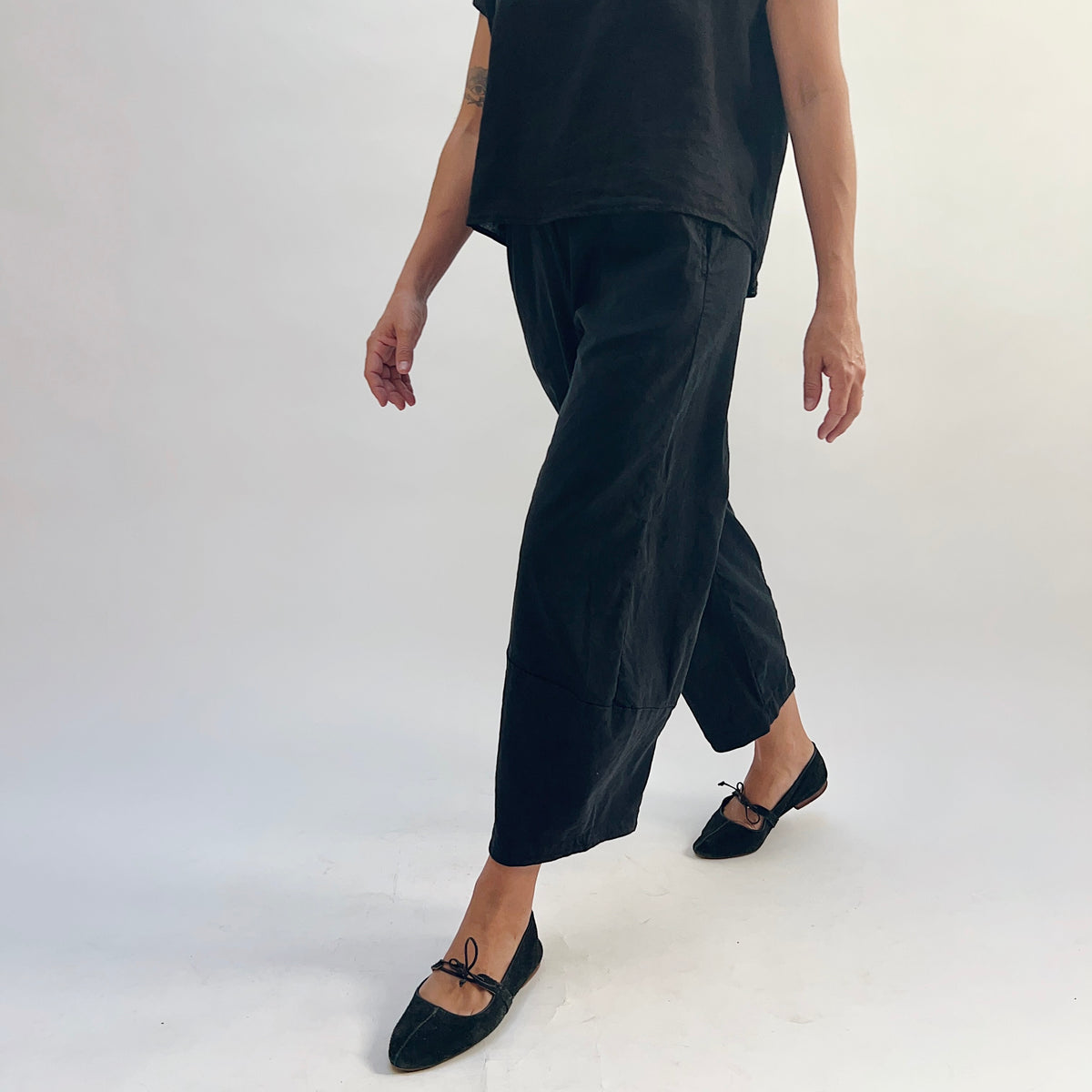 Walking front-angle view of black linen-tencel lantern pants showing wide curved leg silhouette in motion, cropped ankle length and curved hem seam, paired with a black linen top and black bow-tie ballet flats
