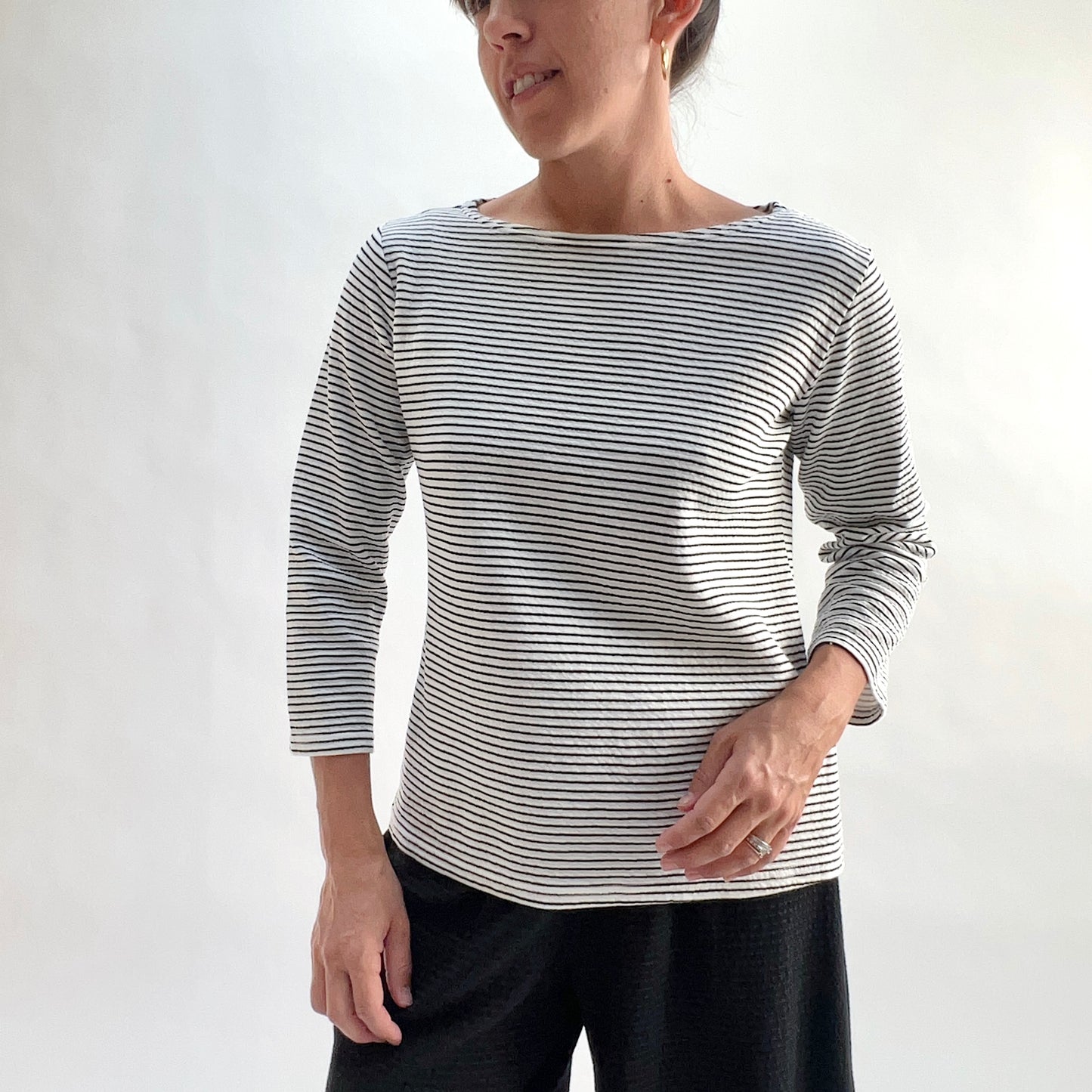 Full front view of model wearing a white and black horizontal stripe boatneck tee with 3/4 sleeves, hand at side, showing the relaxed fit, wide boatneck, and laundered crinkle-texture stripe, styled with black wide-leg pants against a white background.
