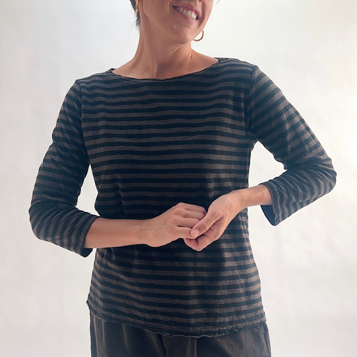 Smiling close front view of model wearing a chocolate and black horizontal stripe boatneck tee with 3/4 sleeves, hands clasped at waist, showing the wide boatneck neckline and linen-blend texture.

