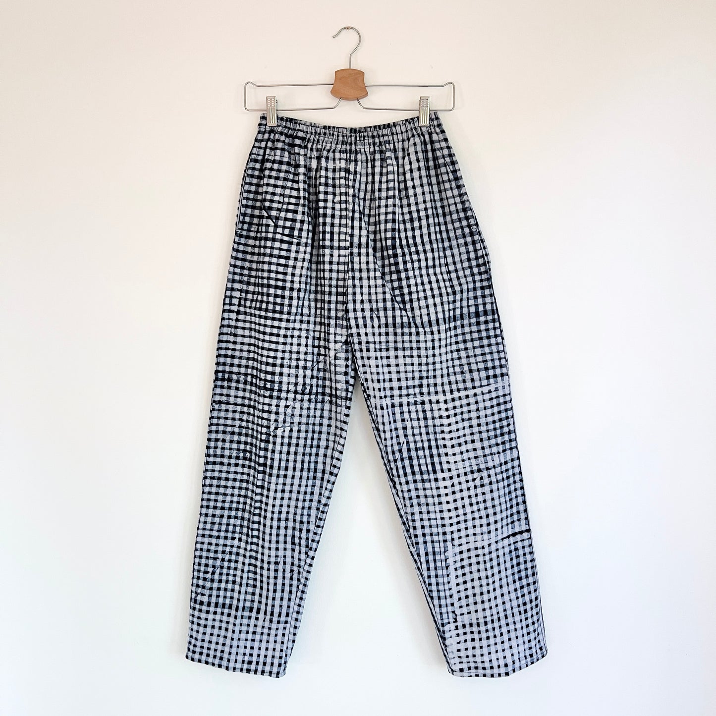 Full front view of Lazy Pants in Indigo Gingham Batiki cotton, hanging on a clip hanger against a white wall, showing elastic waistband, hand-printed black and white gingham check pattern, and relaxed wide-leg silhouette

