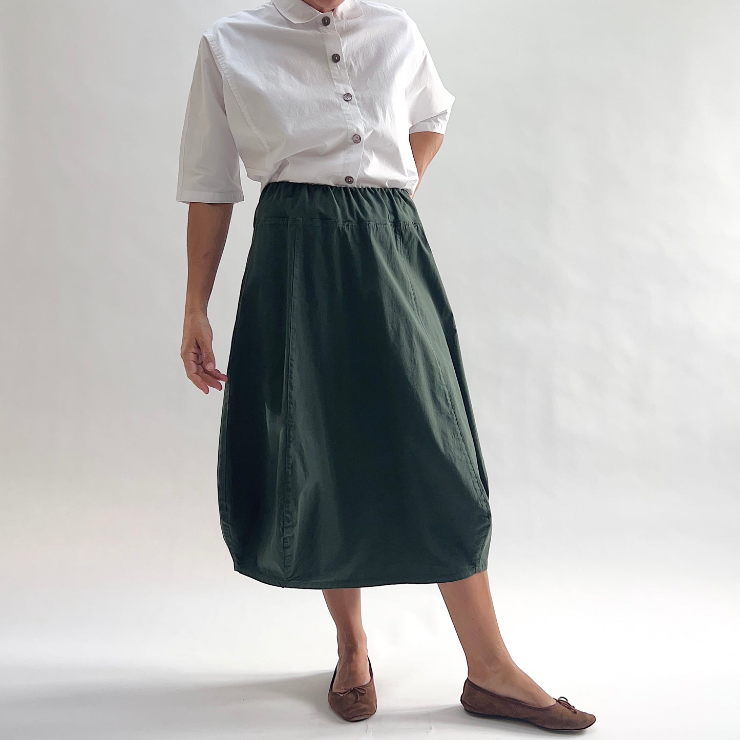 Full front view of model wearing Eleven Stitch konbu rib yoke skirt with arm at side, showing elastic knit waistband, curved seam paneling, sculptural bubble hem, below-the-knee length, and deep forest green color, styled with white chalk camp shirt and brown ballet flats