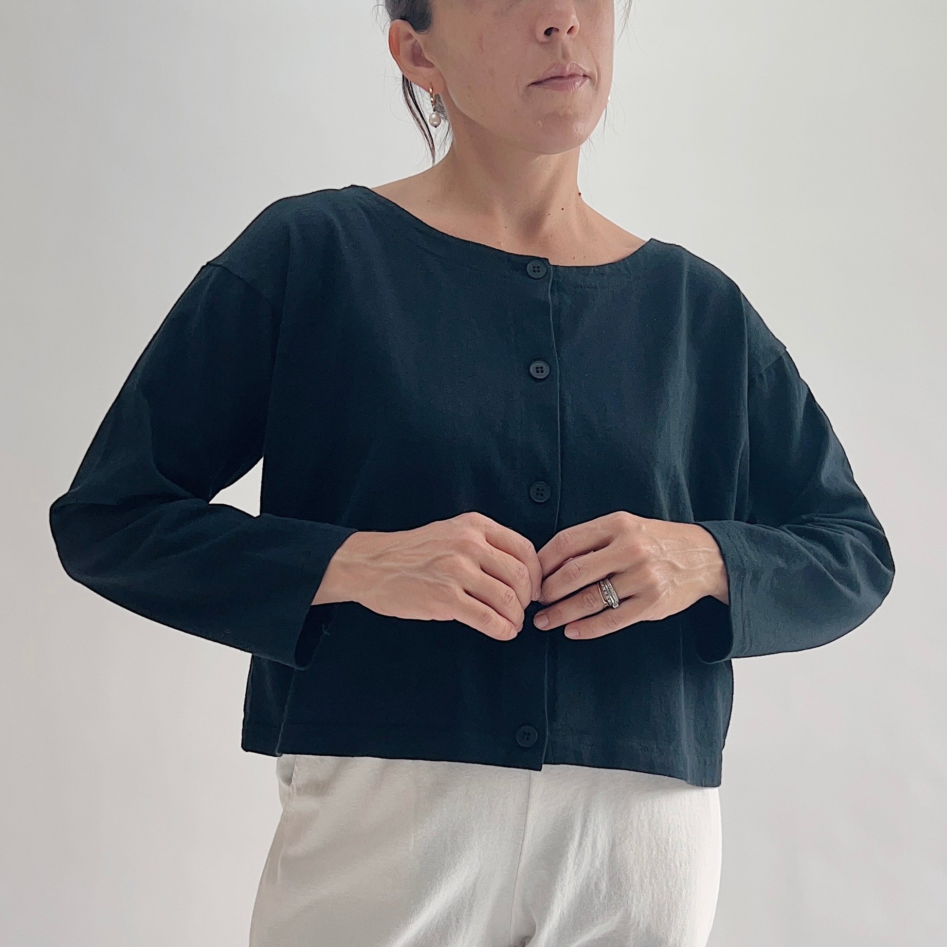 Close front view of model wearing Pacific Cotton black long sleeve button front shirt with hands at buttons, showing wide scoop neckline, five-button center placket, long sleeves, boxy cropped fit, and black color