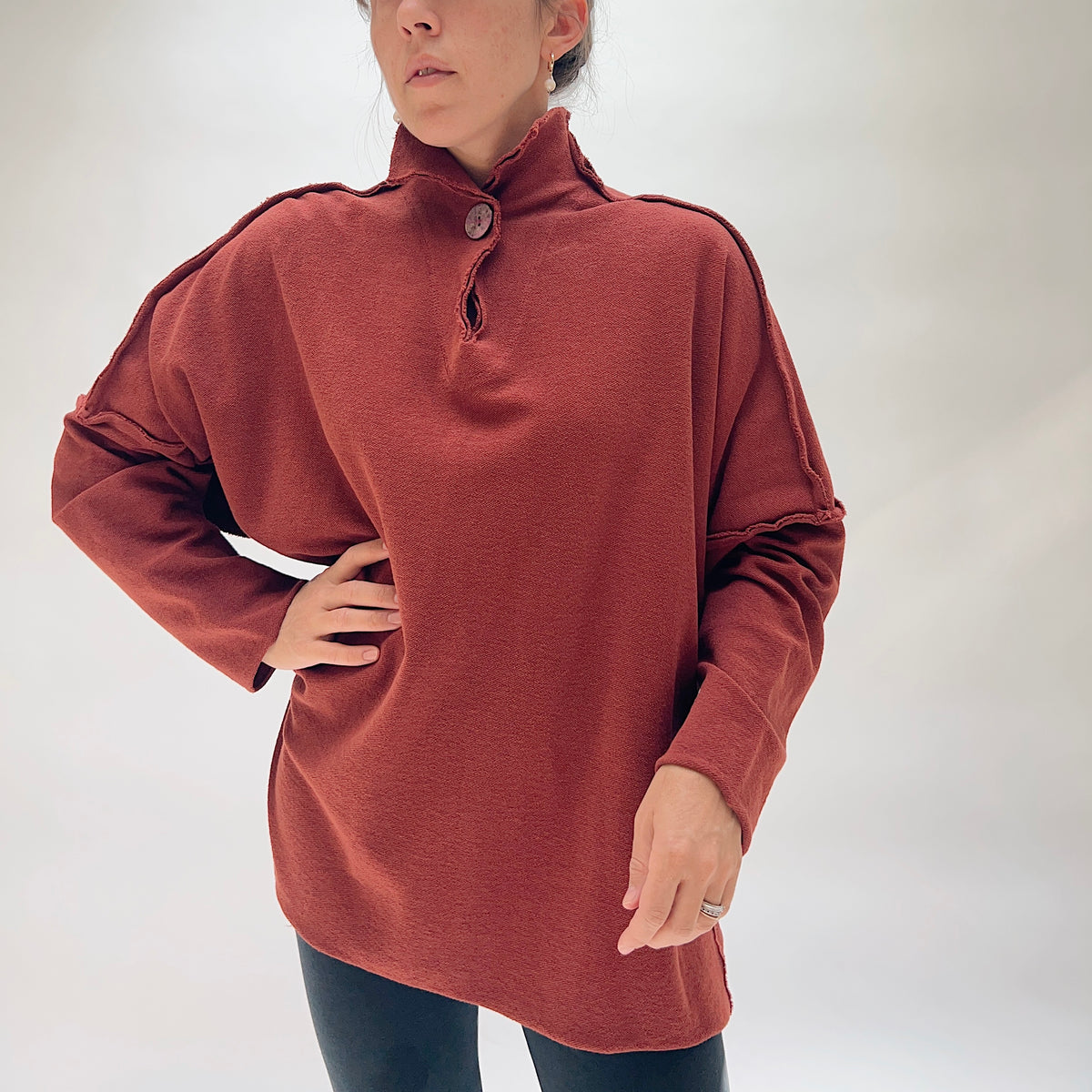 Model wearing the Eleven Stitch Long Funnel Neck Slub Sweatshirt in Ginger Spice from the front — a tunic-length oversized top in warm terracotta slub cotton with a button funnel neckline, exposed raw seam detailing at the shoulders, drop sleeves, and a relaxed cocoon silhouette, paired with dark jeans
