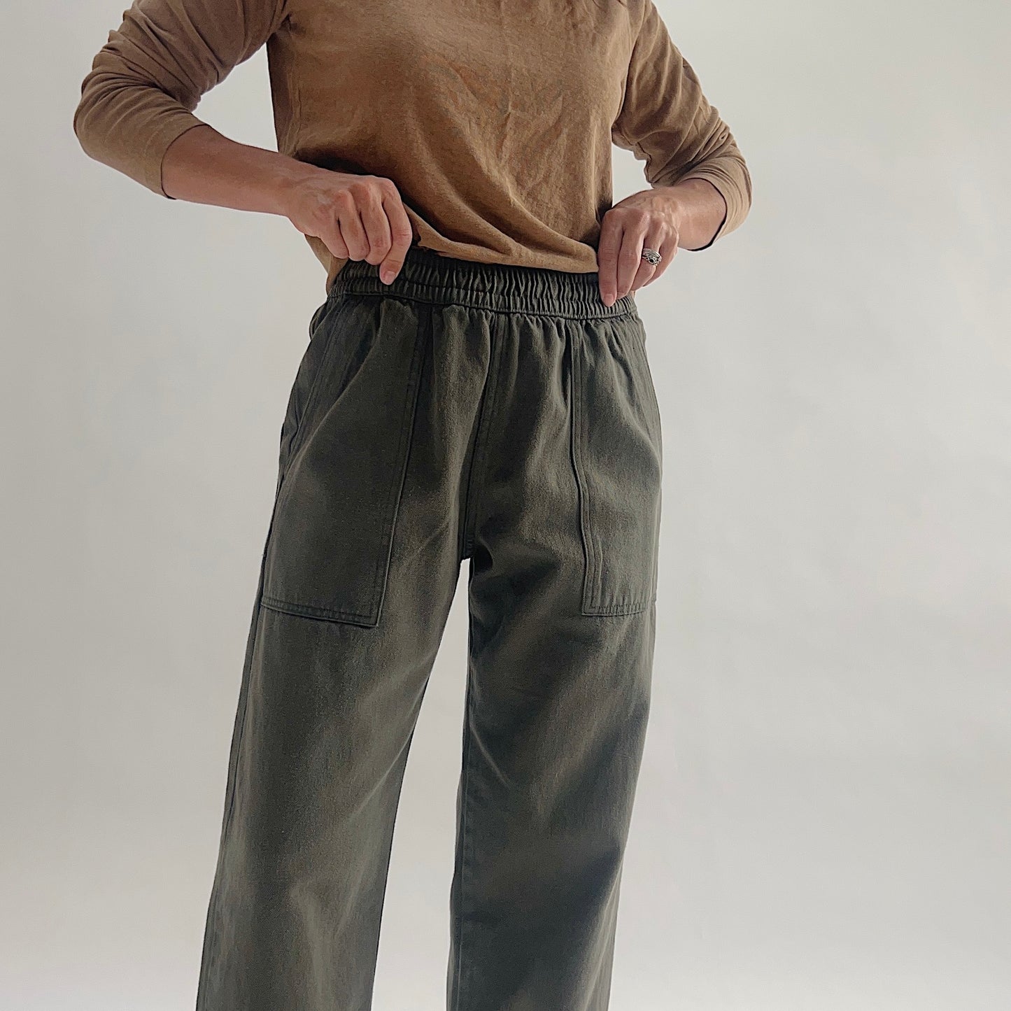 Close-up of the elastic waist and front patch pockets on olive green wide-leg pull-on pants, with model tucking in a tan long-sleeve top.
