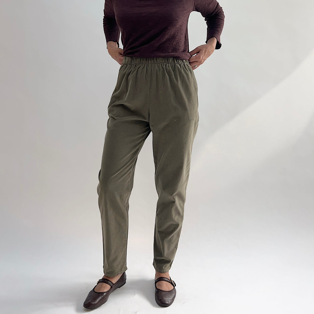 Model wearing the Bryn Walker Corduroy Boslow Pants in Jasper, front view showing fully elastic waistband, side pockets, relaxed tapered leg, and ankle length in olive jasper pinwheel corduroy, styled with burgundy Double Cotton Tee and brown Mary Jane flats
