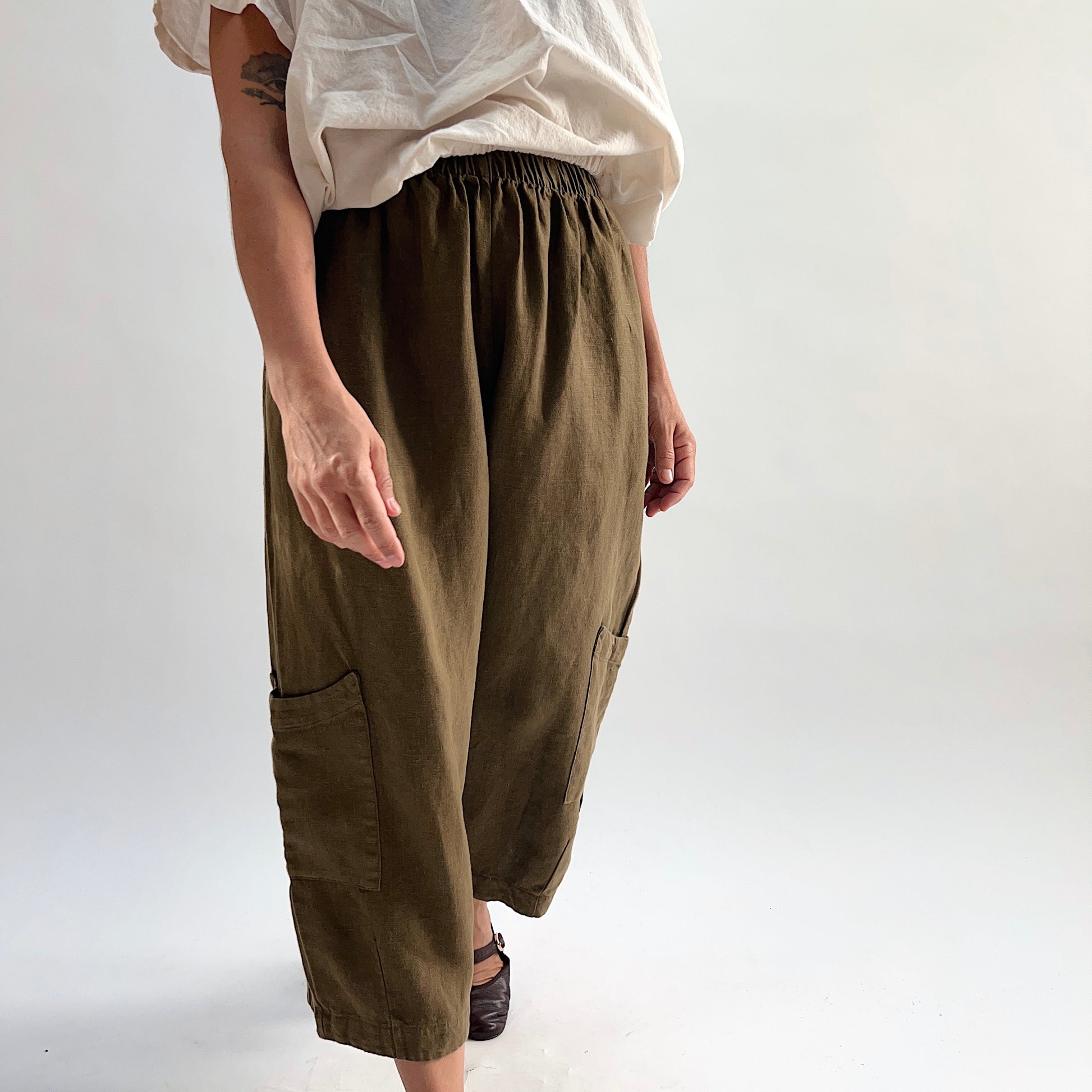 Side view of the Bryn Walker Heavy Linen Casbah Pant in Partridge, highlighting elastic waistband gathers, patch pocket at lower leg, and drape of olive heavyweight linen, with dark Mary Jane flats
