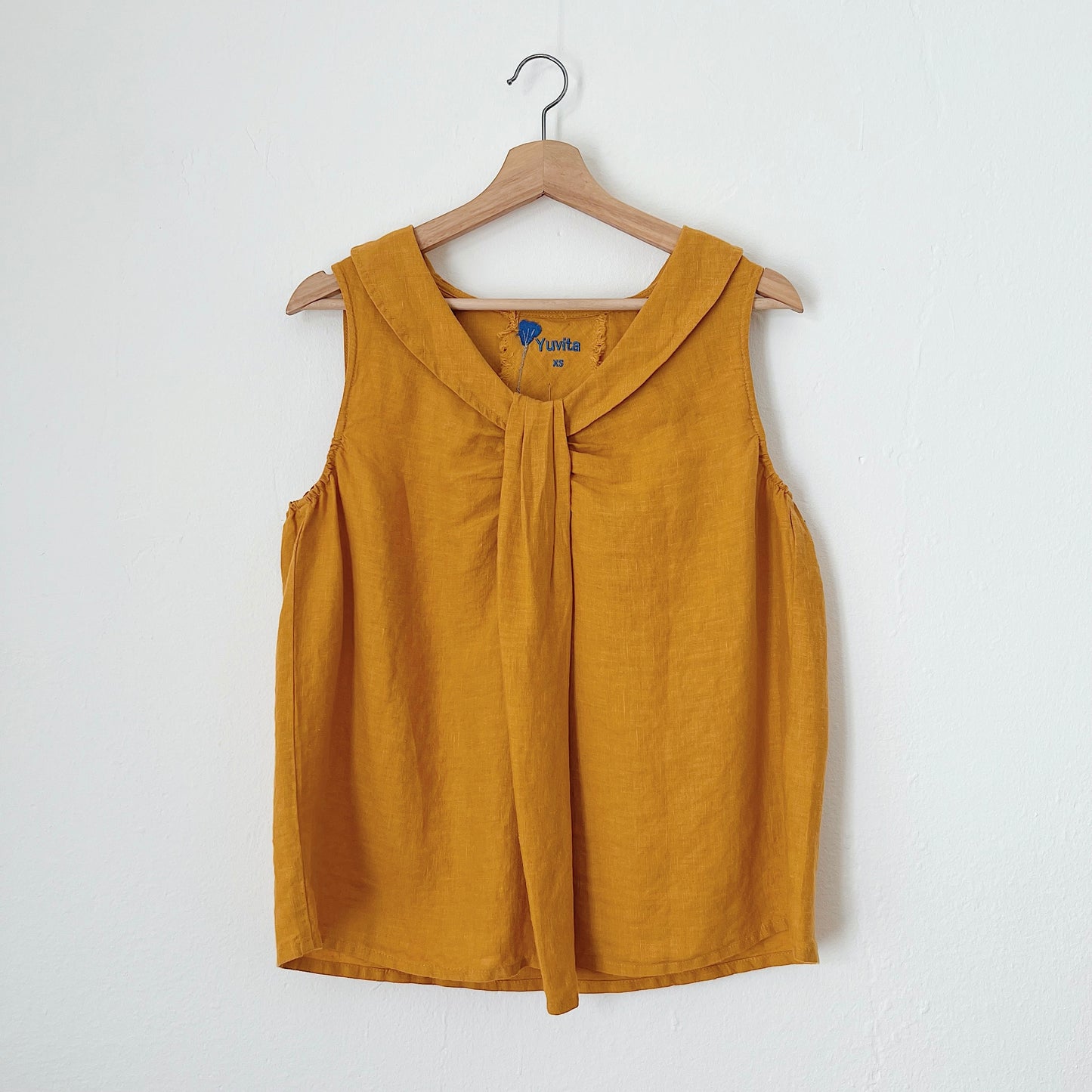 A sleeveless, mustard yellow linen top hanging on a wooden hanger against a white background.