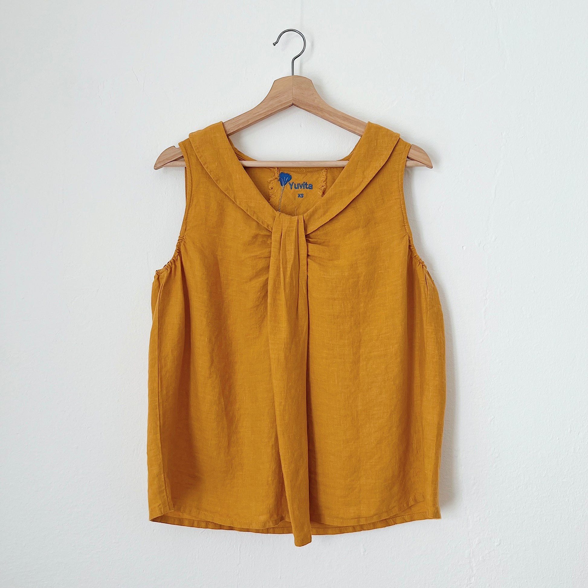 A sleeveless, mustard yellow linen top hanging on a wooden hanger against a white background.