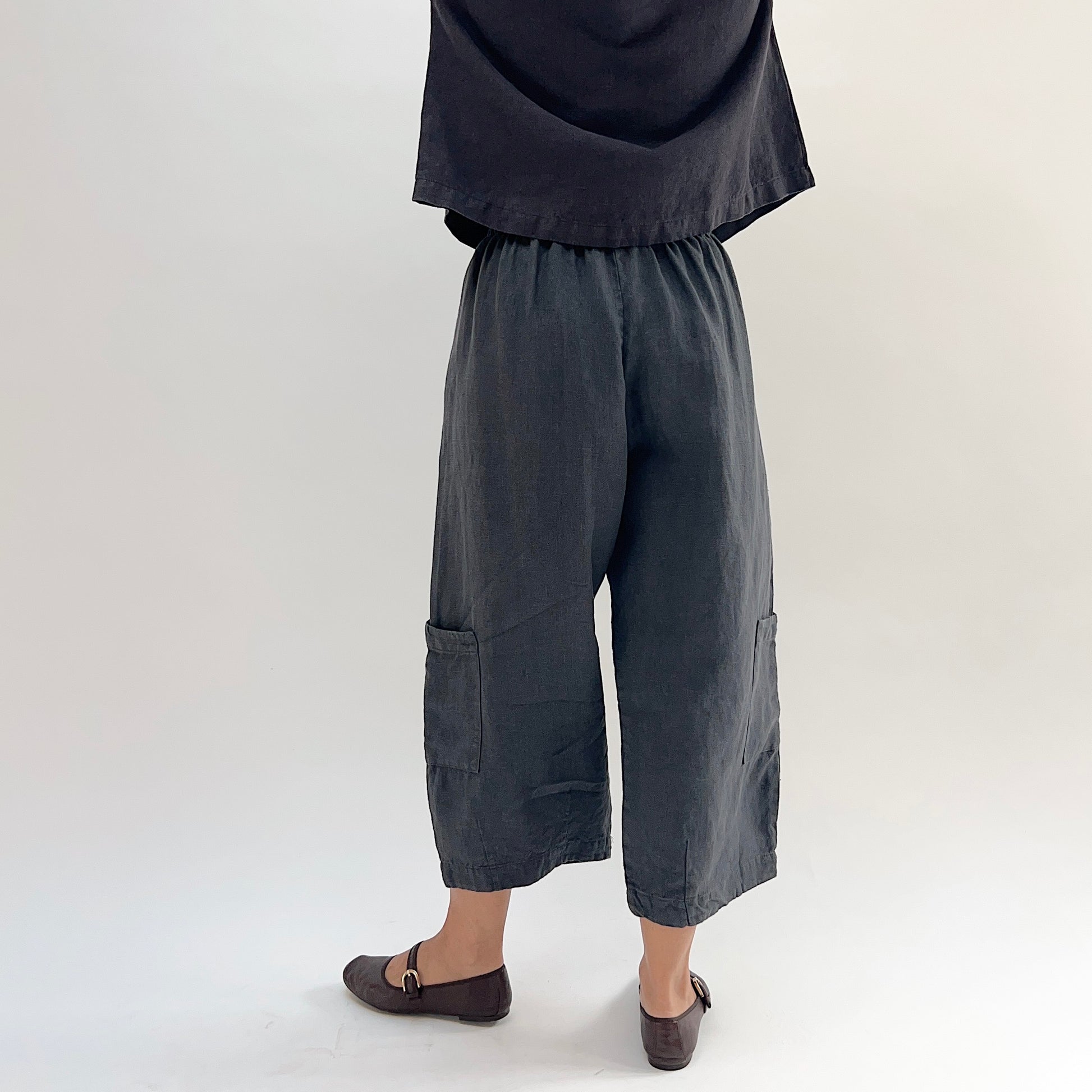 Back view of model wearing the Bryn Walker Heavy Linen Casbah Pant in Moonlight, showing fully elastic waistband, barrel-tapered leg, two large lower-leg patch pockets, cropped ankle length, and deep moonlight heavyweight linen, styled with matching linen top and brown Mary Jane flats
