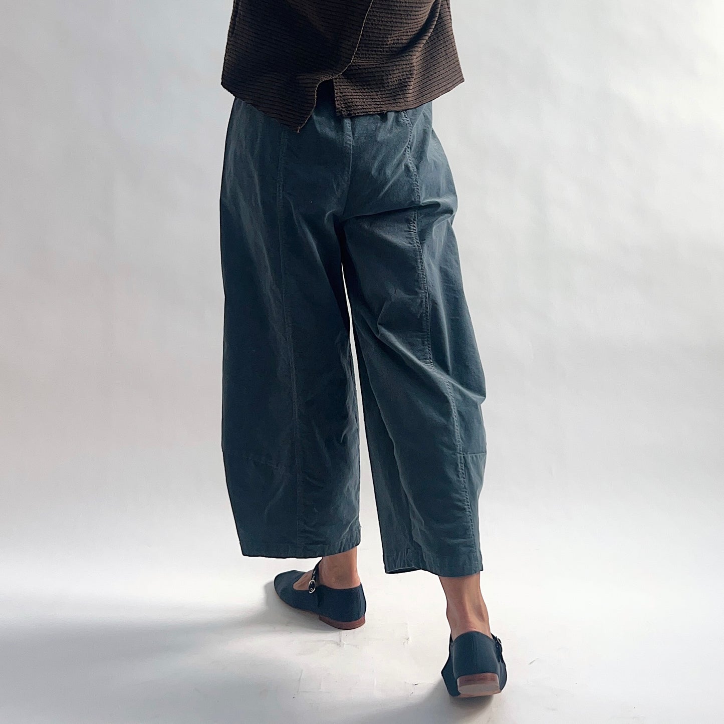 Back view of model wearing iron blue-gray mini cord lantern pants, showing the elastic waist gathering, side seam, wide rounded volume, and tapered ankle, styled with the matching chocolate striped cardigan and blue mary jane flats against a white background.
