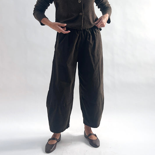 Full front view of model wearing chocolate mini cord lantern pants with elastic gathered waist, hands on hips, showing the wide rounded silhouette and tapered ankle, styled with the matching striped cropped cardigan and dark mary jane flats against a white background.
