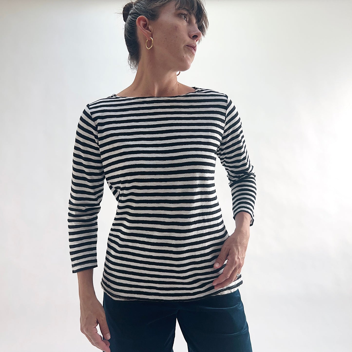 Three-quarter front view of model wearing a navy and white laundered stripe 3/4 sleeve boatneck tee, looking to the side with hand on hip, showing the boatneck neckline and relaxed silhouette, styled with dark blue pants.
