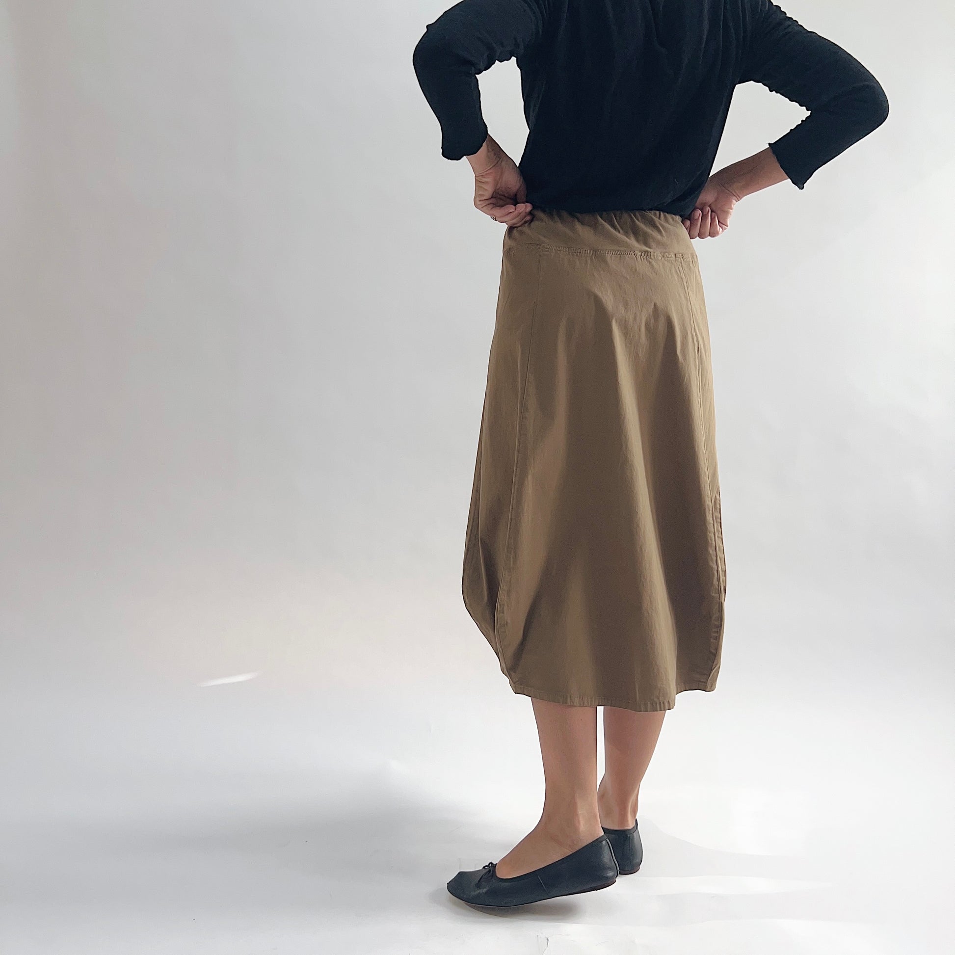 Back view of model wearing Eleven Stitch winter olive rib yoke skirt with hands on hips, showing elastic knit waistband, curved seam paneling, sculptural bubble hem, below-the-knee length, and warm khaki olive color, styled with black long sleeve top and black ballet flats