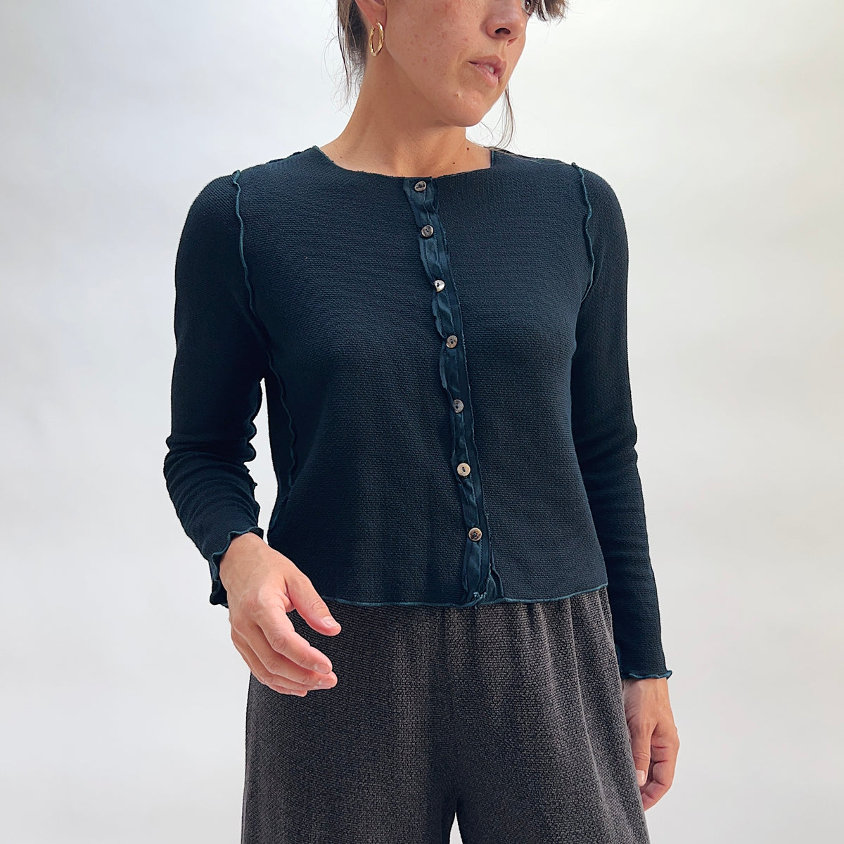 Model wearing the Cut Loose Marled Knit Cropped Cardigan in Etan, three-quarter front view, showing full button front with iridescent shell buttons, scalloped lettuce edges at neckline and hem, and cropped silhouette of deep teal marled knit, styled with a dark textured skirt
