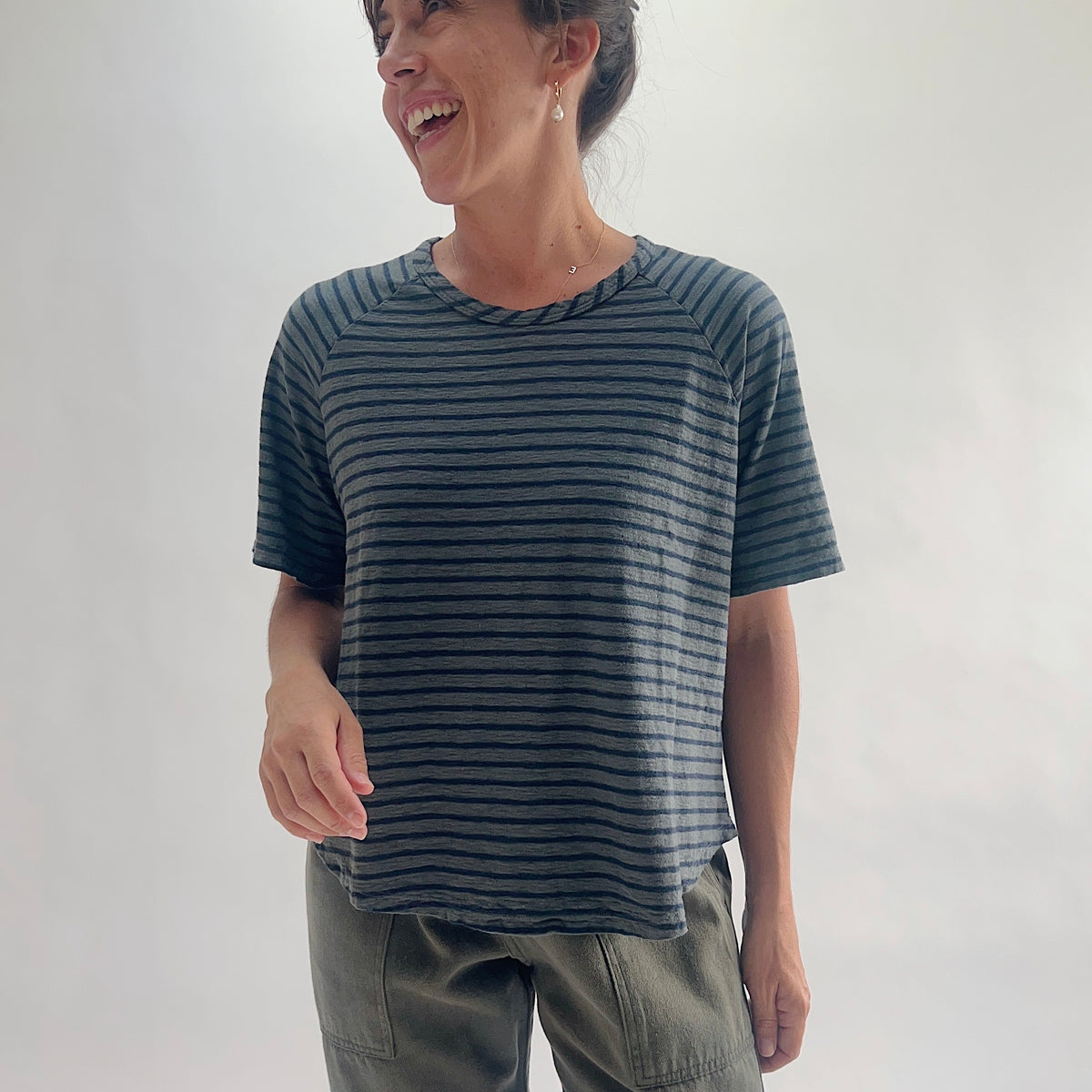 Full front view of model smiling in an anthracite and navy striped boxy raglan tee with a relaxed boxy silhouette and curved hem, styled with olive green pants.
