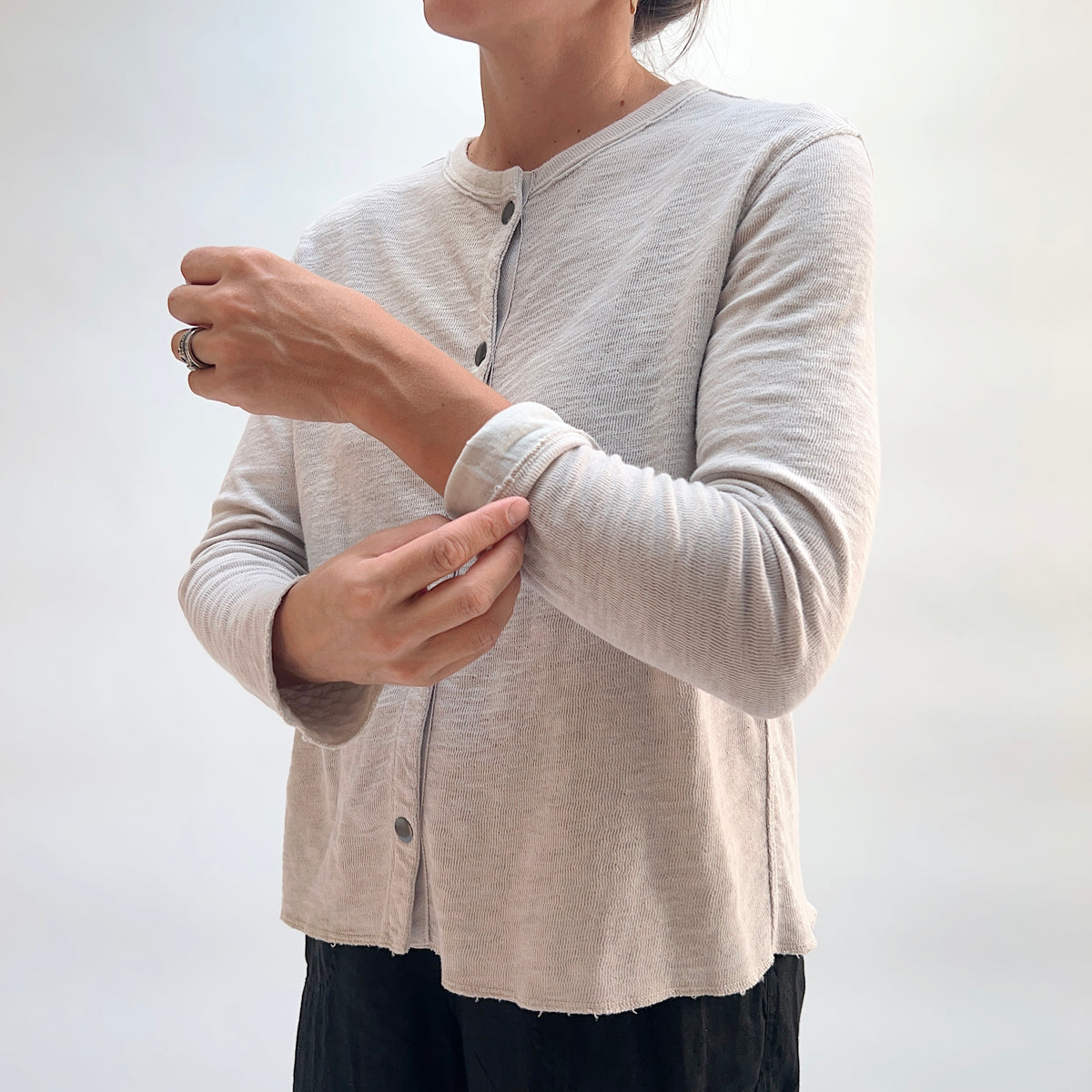 Close-up of model adjusting the sleeve of the Cut Loose Snap Front Cardigan in Nimbus, highlighting snap button closure and raw hem detail in light heathered gray cotton
