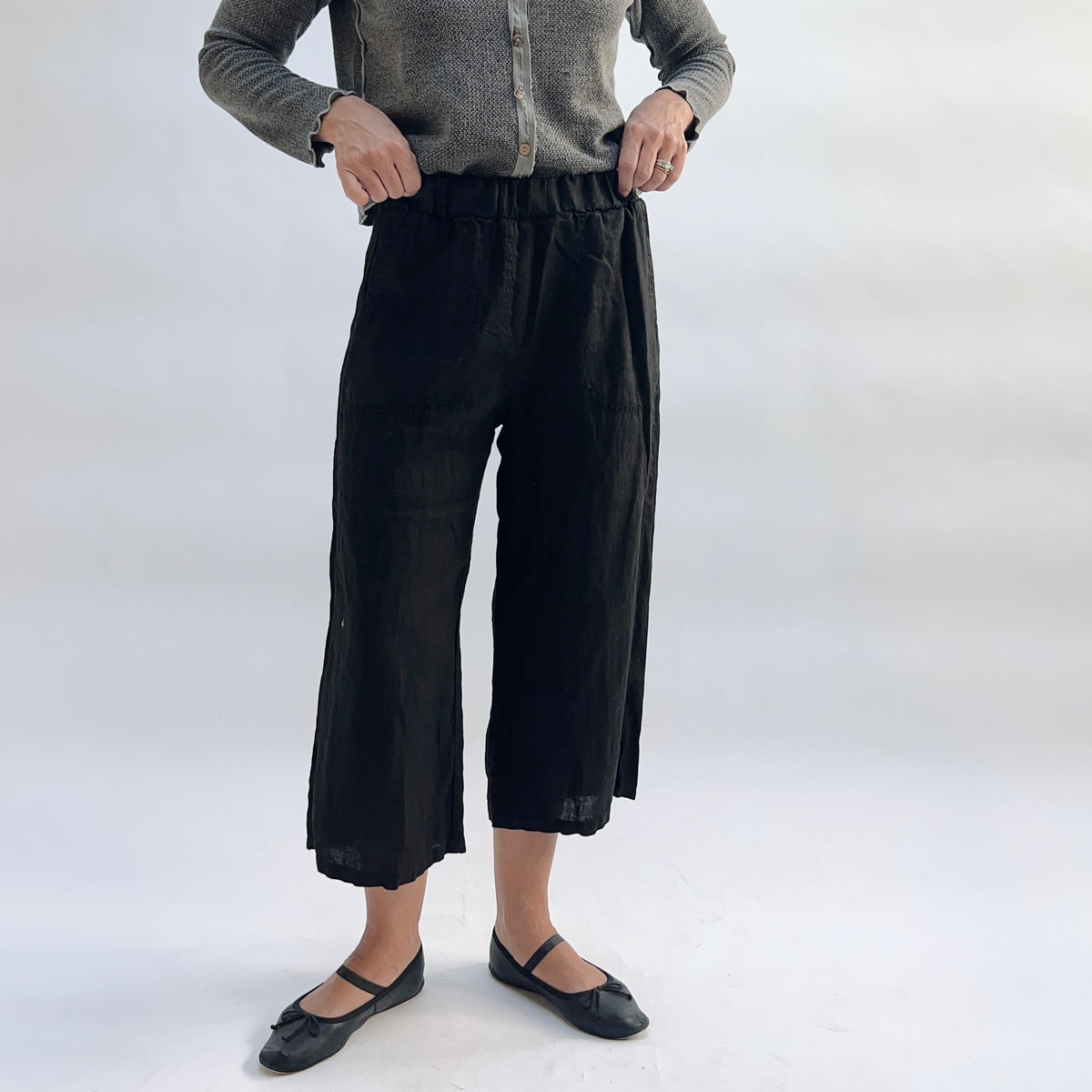 Cut Loose Hanky Linen Easy Crop Flood Pant in Black, black linen wide-leg crop pant with hands pulling at elastic waistband, styled with a grey button-front cardigan and black Mary Jane flats — front view showing elastic waist and patch pocket detail
