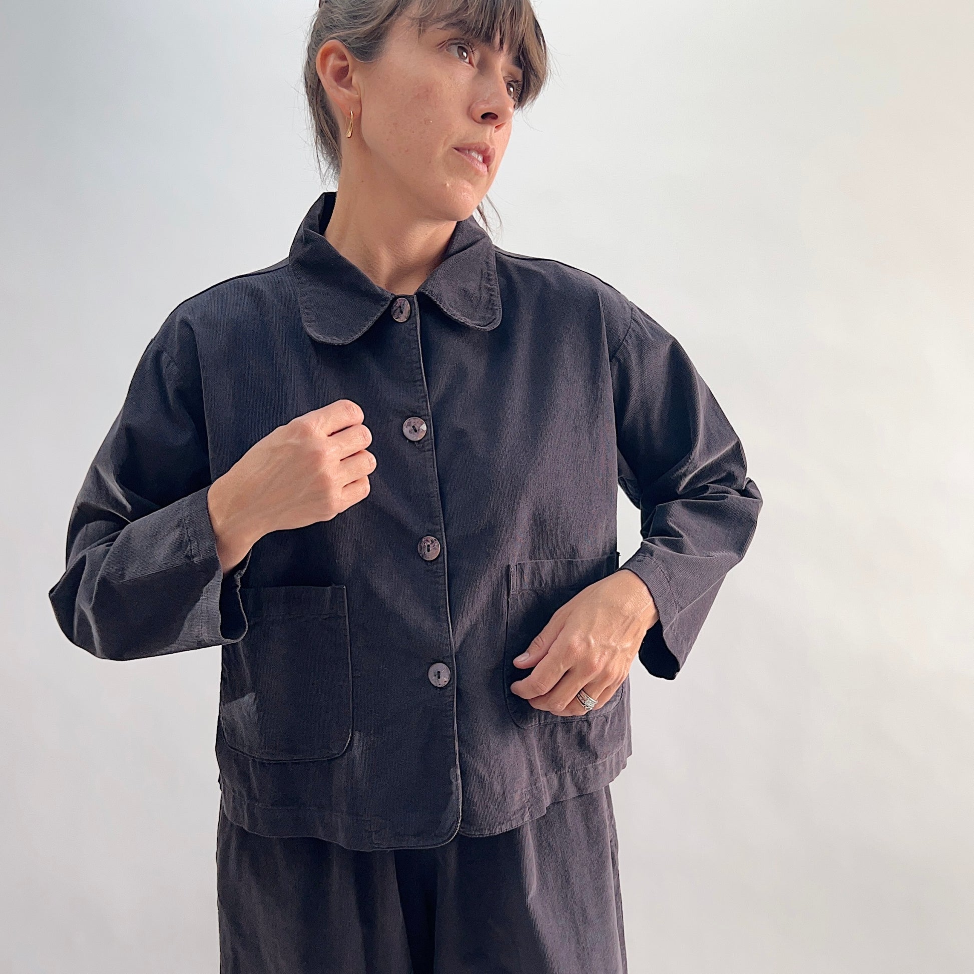 Model adjusting lapel of the Bryn Walker Corduroy Audrey Jacket in Fig, close-up front view showing rounded collar, shell buttons, and patch pockets in deep charcoal corduroy
